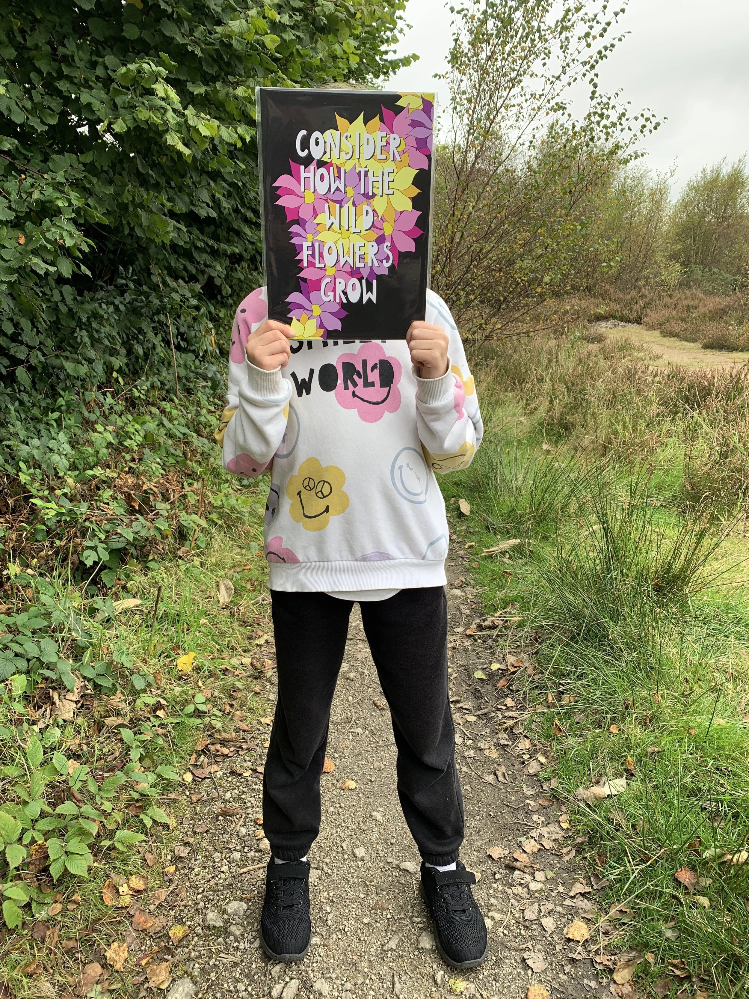 Person standing on a dirt trail surrounded by green foliage, wearing a white sweatshirt decorated with smiley and flower prints, black pants, and black shoes, holding a sign with colorful flowers and the text 'Consider how the wild flowers grow'
