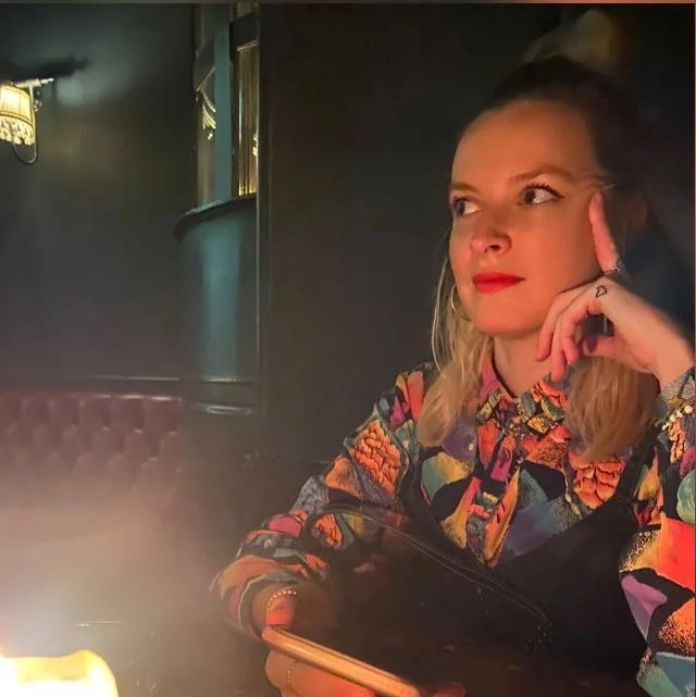 A woman with blonde hair and red lipstick sitting at a table in a dimly lit restaurant or cafe, wearing a colorful floral patterned top, with her right hand resting on her face and looking thoughtfully to the side.
