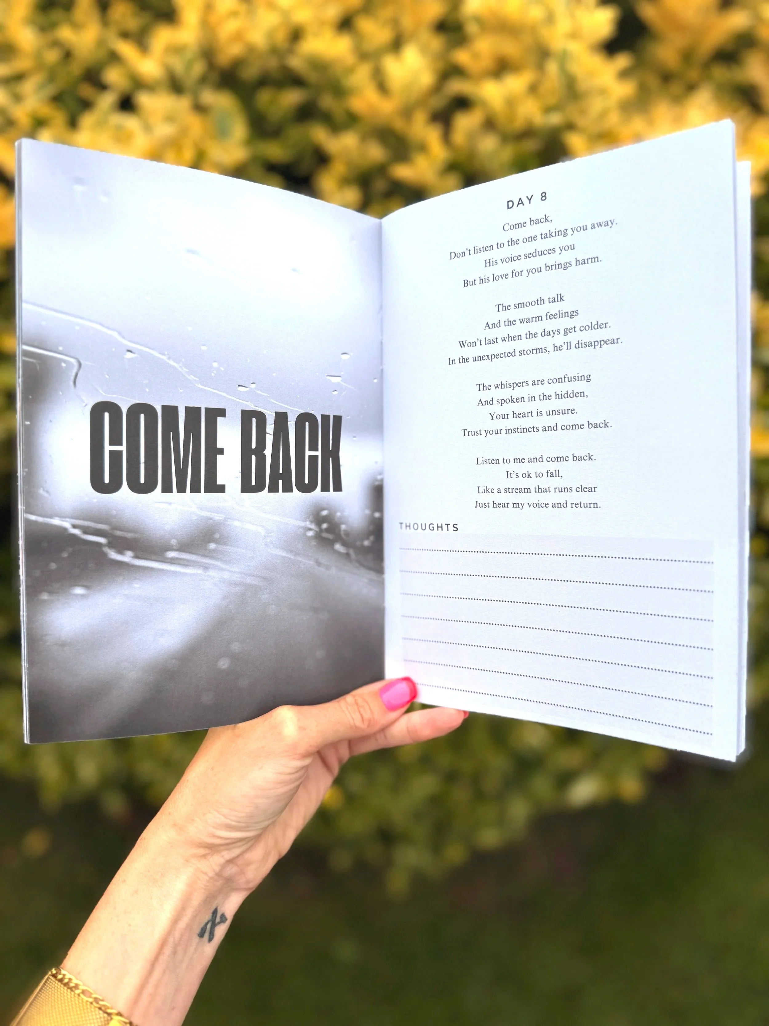 Open journal or planner with the words 'COME BACK' on the left page and a poem titled 'Day 8' on the right page. The background is blurred foliage, and a person with pink nail polish and a gold bracelet is holding the journal.