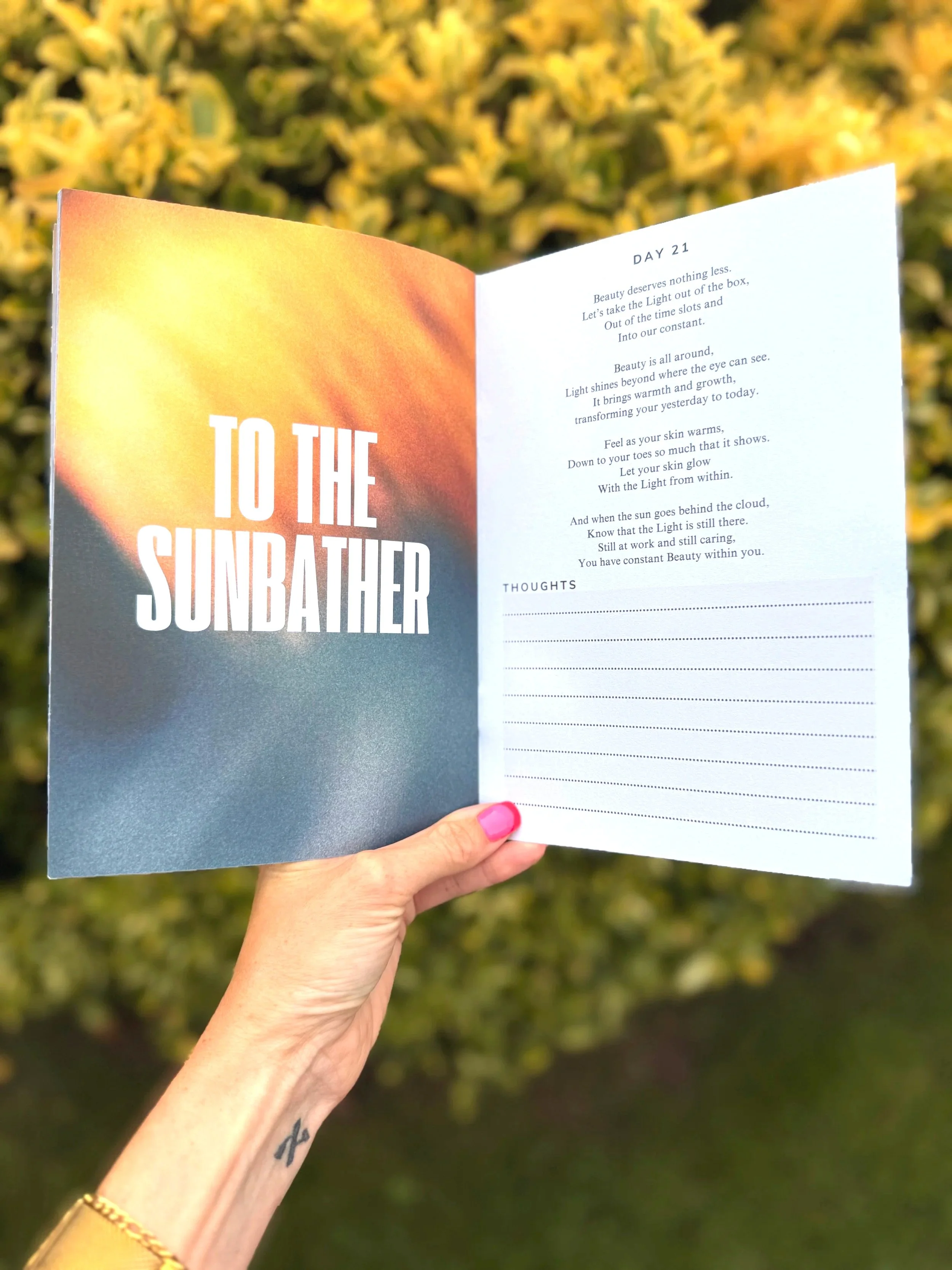 Open book titled 'To the Sunbath' with a sunset background on the left page and a poem titled 'Day 21' on the right page, outdoors with green foliage in the background.