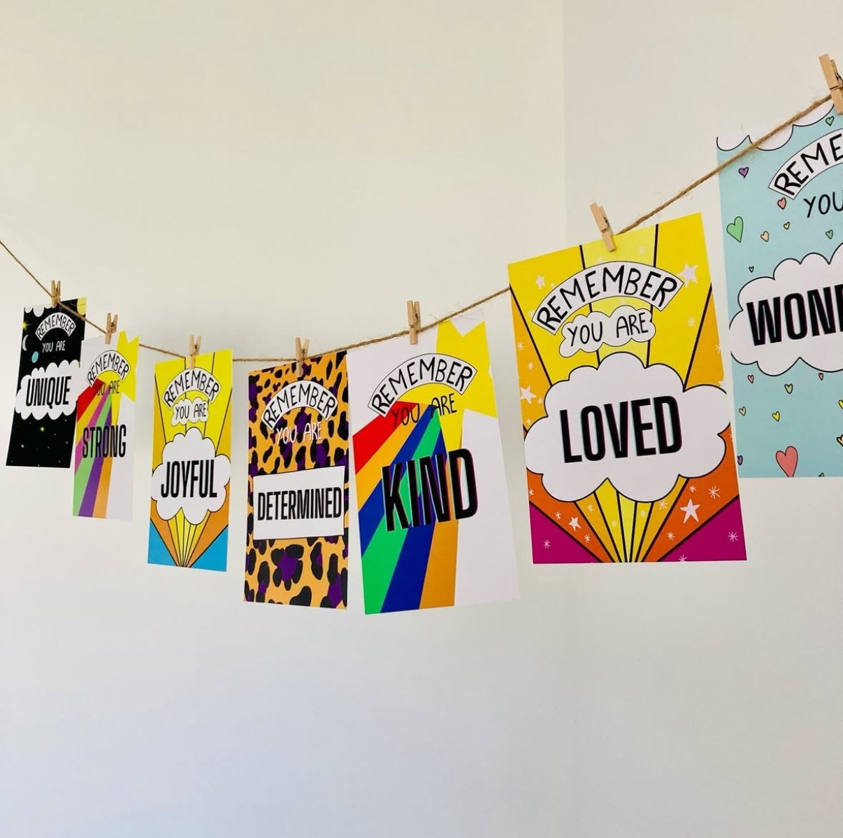 Colorful posters hanging on a string with clothespins, each with positive affirmations like 'Remember you are loved,' 'Remember you are kind,' and 'Remember you are unique,' featuring bright rainbow, animal print, and heart designs.