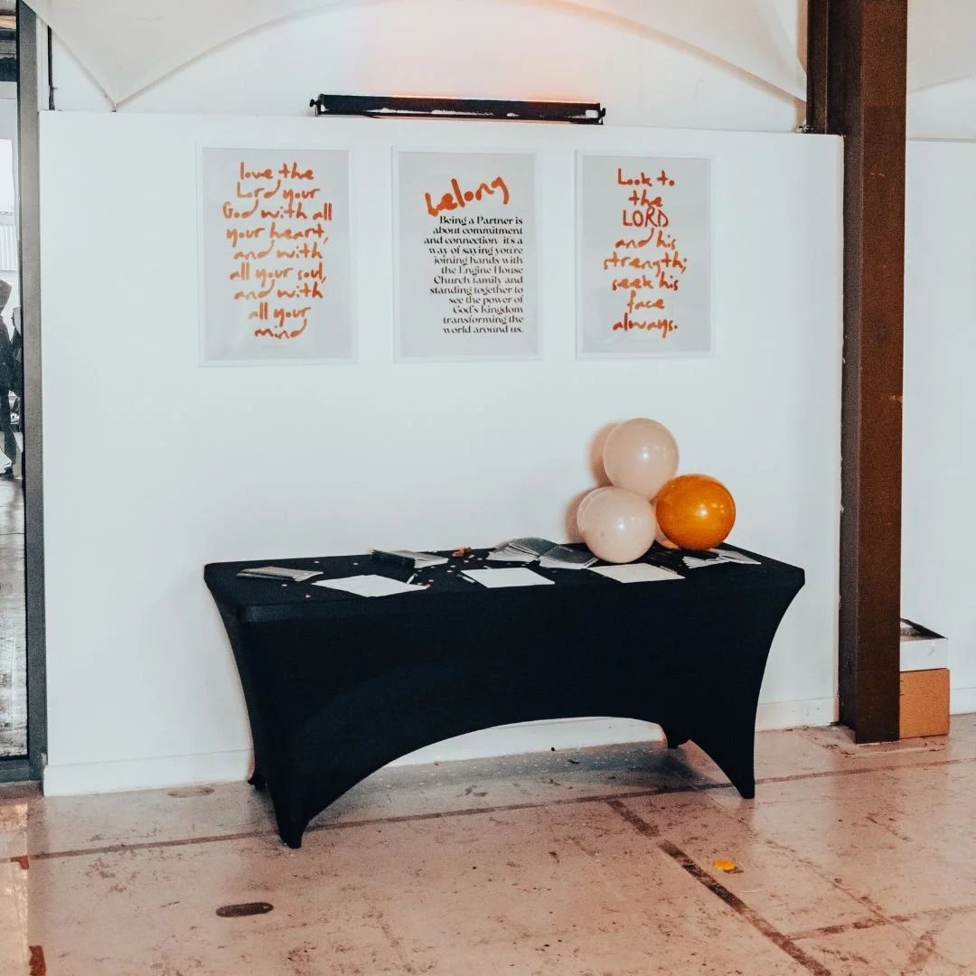 Display table covered with brochures and papers, with three balloons (two pink and one orange) on the right side. Behind the table, there are three framed posters with orange and black text hung on a white wall.
