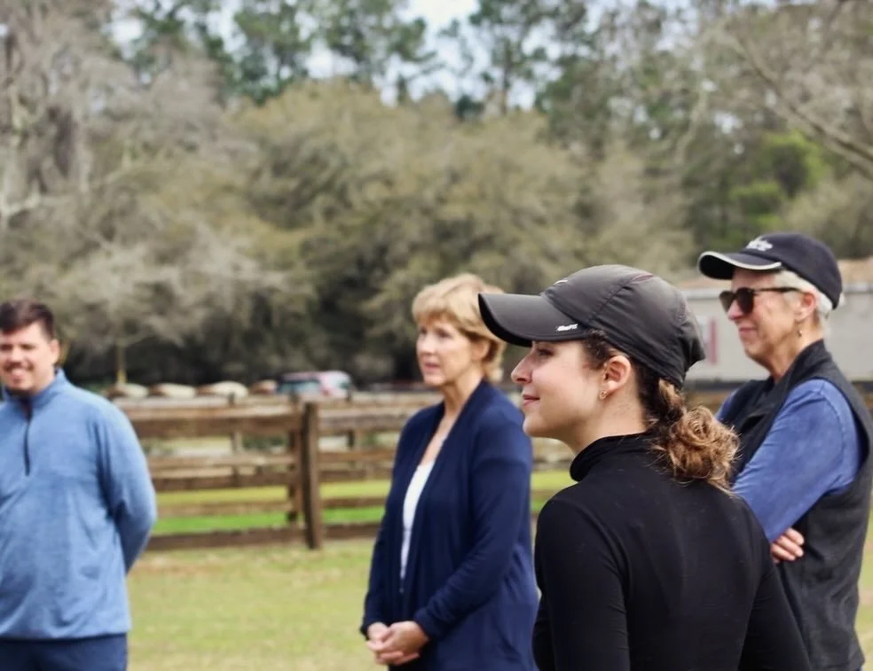 Four people standing outdoors in a field, wearing casual and outdoor clothing, with trees and a wooden fence in the background.