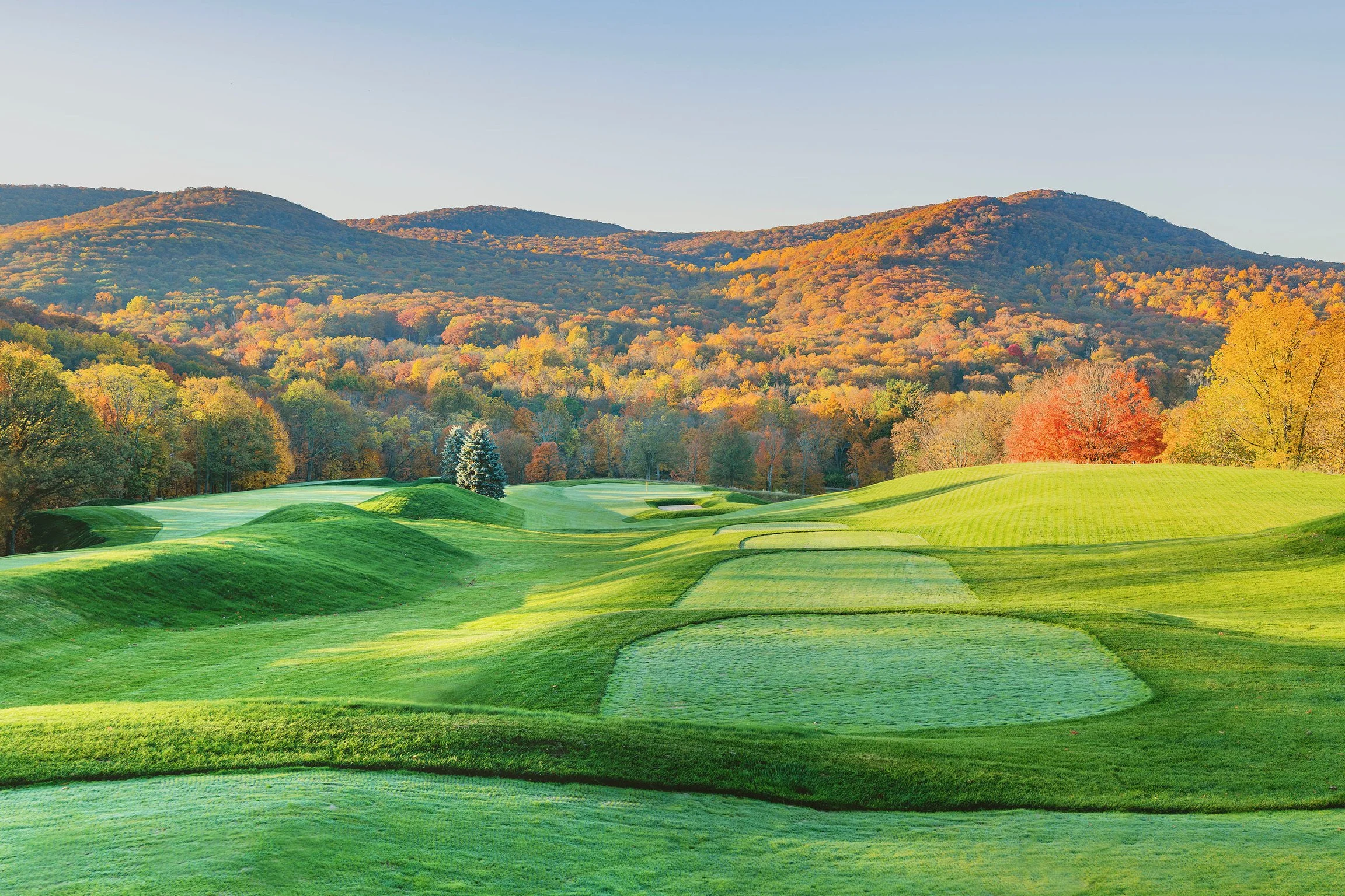 A scenic view of a golf course with neatly mowed greens and sand traps, set against a backdrop of colorful autumn trees and distant mountains under a clear sky.