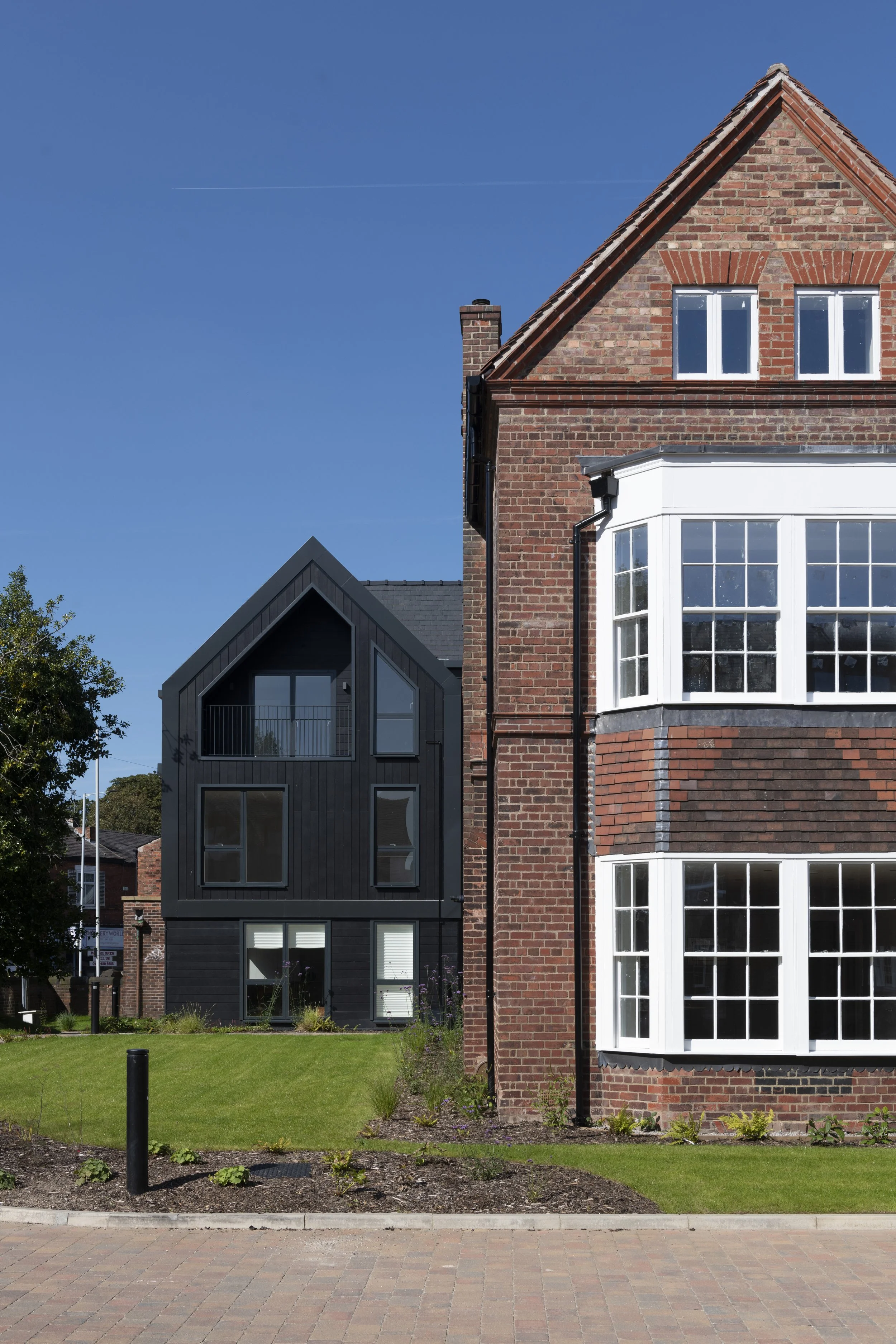 A modern black house with large windows located behind a lush green lawn and flower bed on a sunny day, with other houses and trees visible in the background.