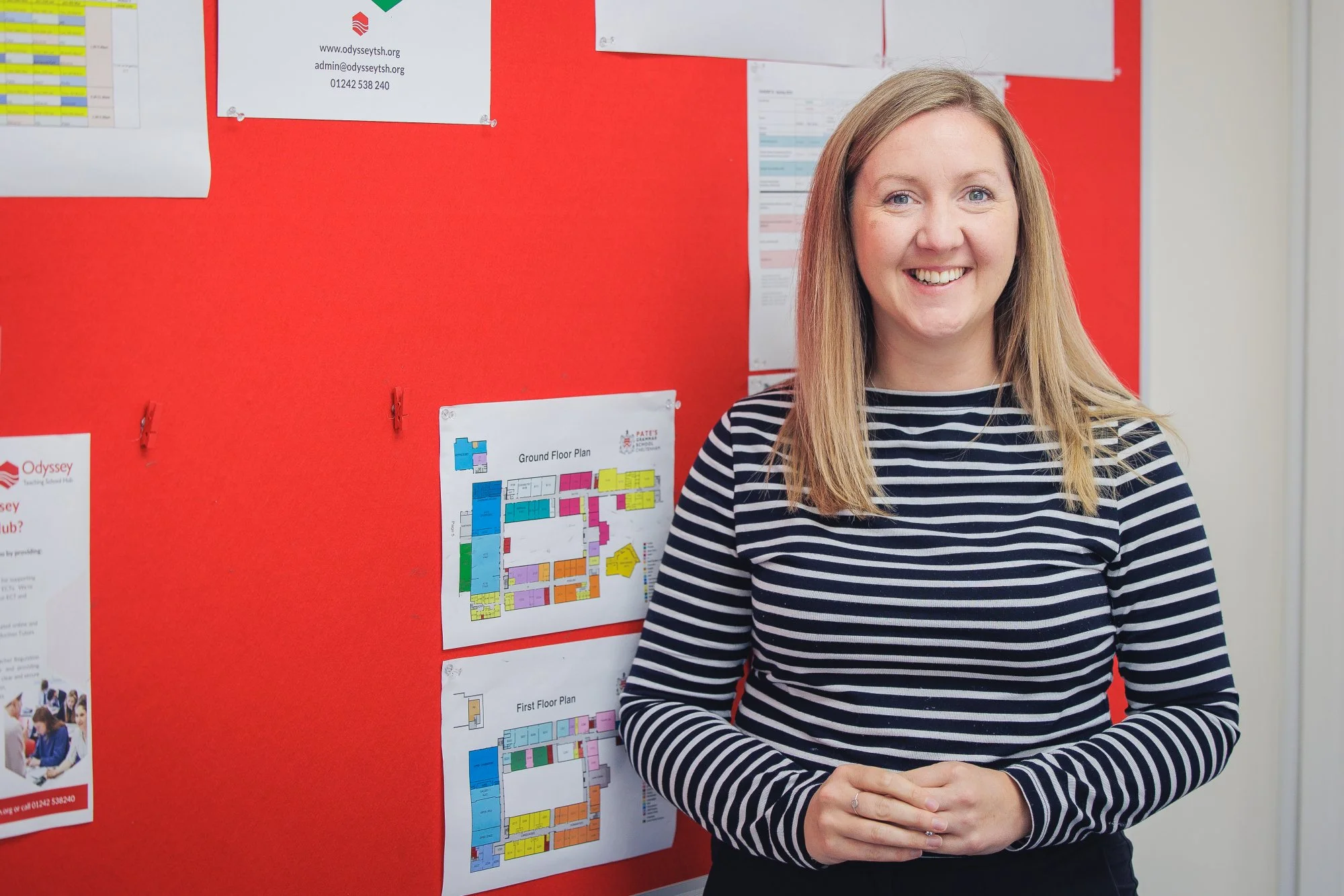A smiling woman with long blonde hair standing in front of a red bulletin board with maps and notices pinned to it.