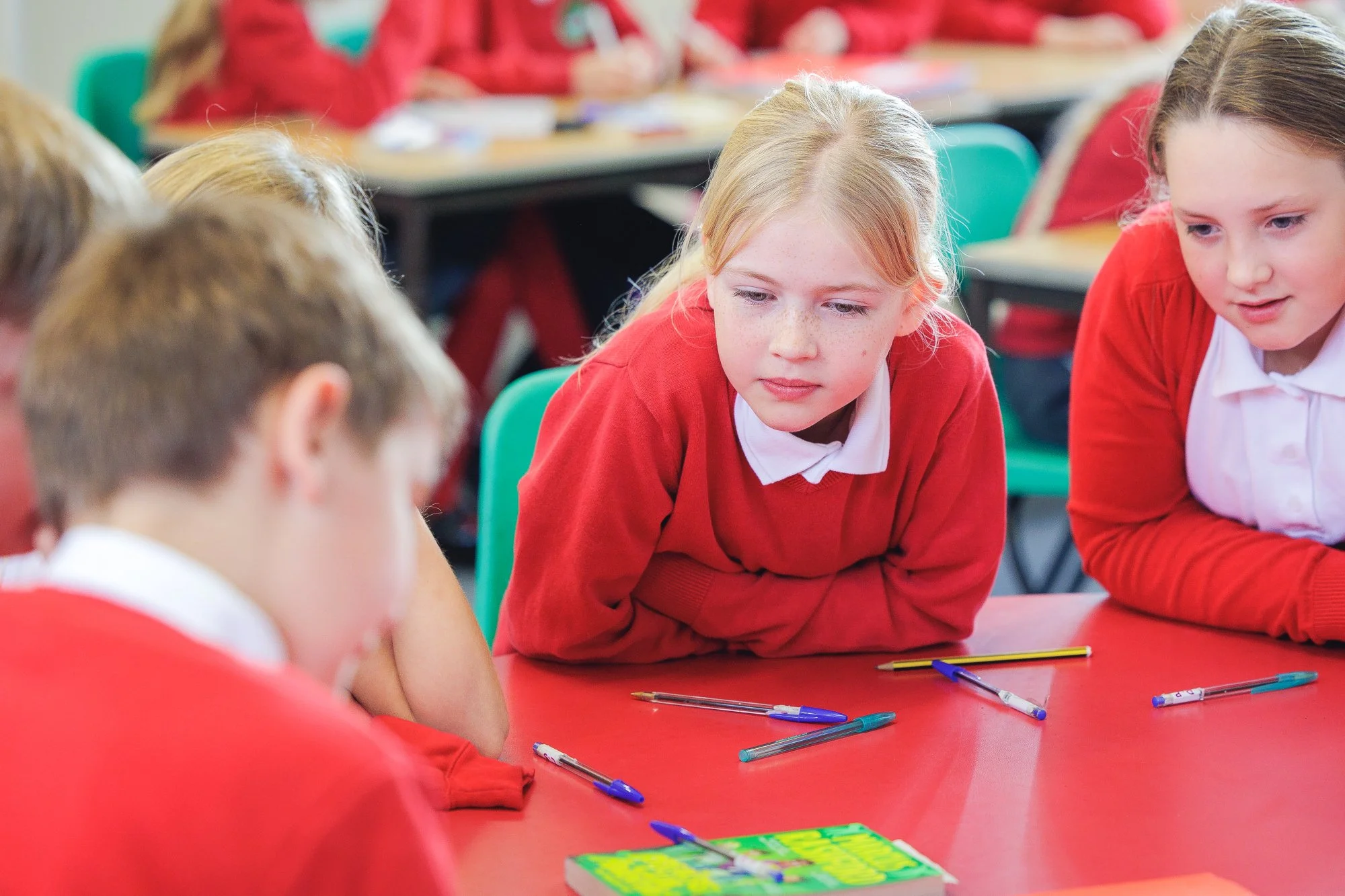 Group of children in school uniforms sitting around a table with pens and a box of snacks, engaged in a discussion.