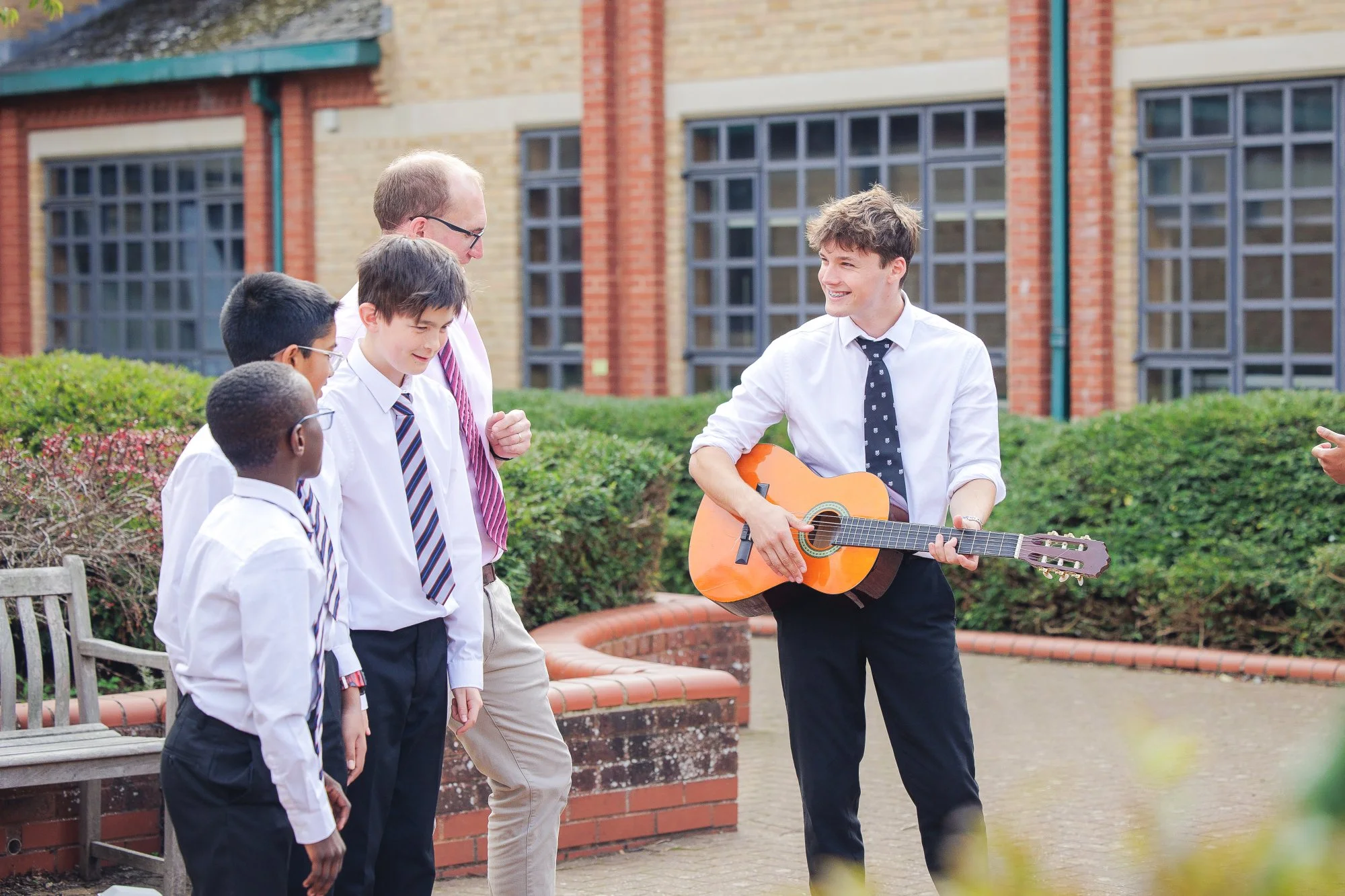 A young man playing guitar and singing to four male students in school uniforms outdoors, with a school building and greenery in the background.