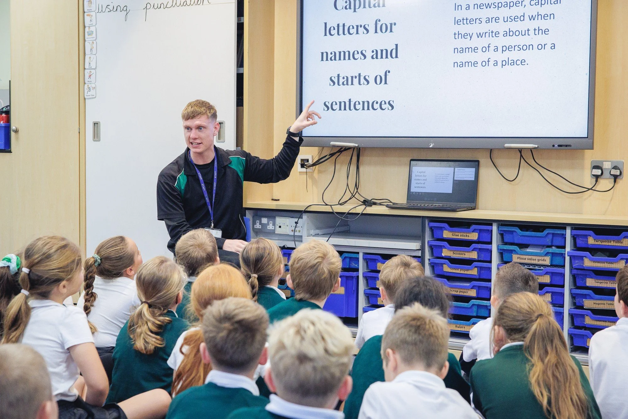 A teacher standing in front of a classroom pointing at a large screen displaying educational content about capital letters, with students sitting on the floor and paying attention.
