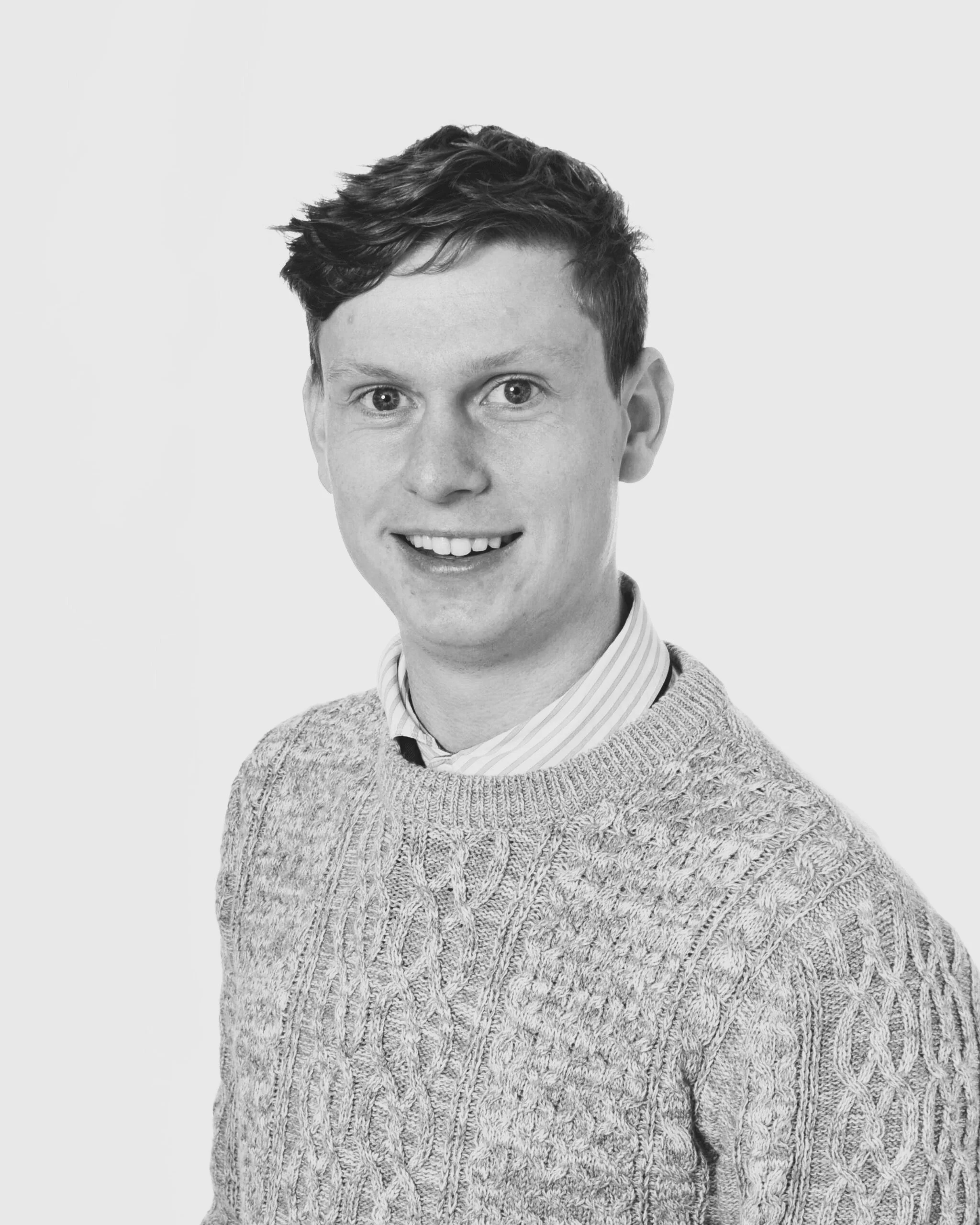 A black and white portrait of a young man with short, wavy hair, smiling at the camera, wearing a patterned knit sweater over a collared shirt.