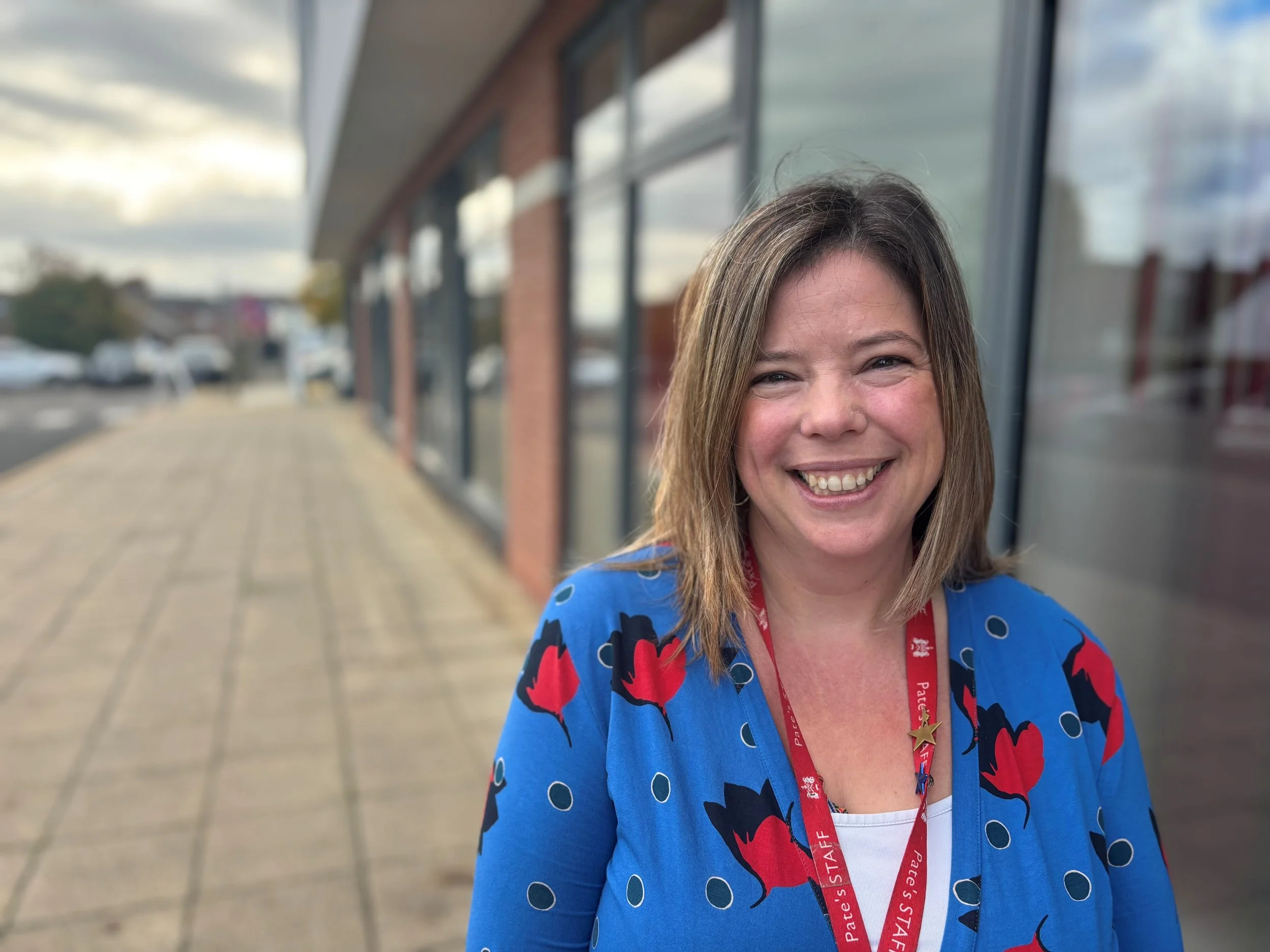 A smiling woman with shoulder-length brown hair wearing a blue jacket with red and black floral patterns, and a red lanyard around her neck, standing outside near a building with large glass windows.