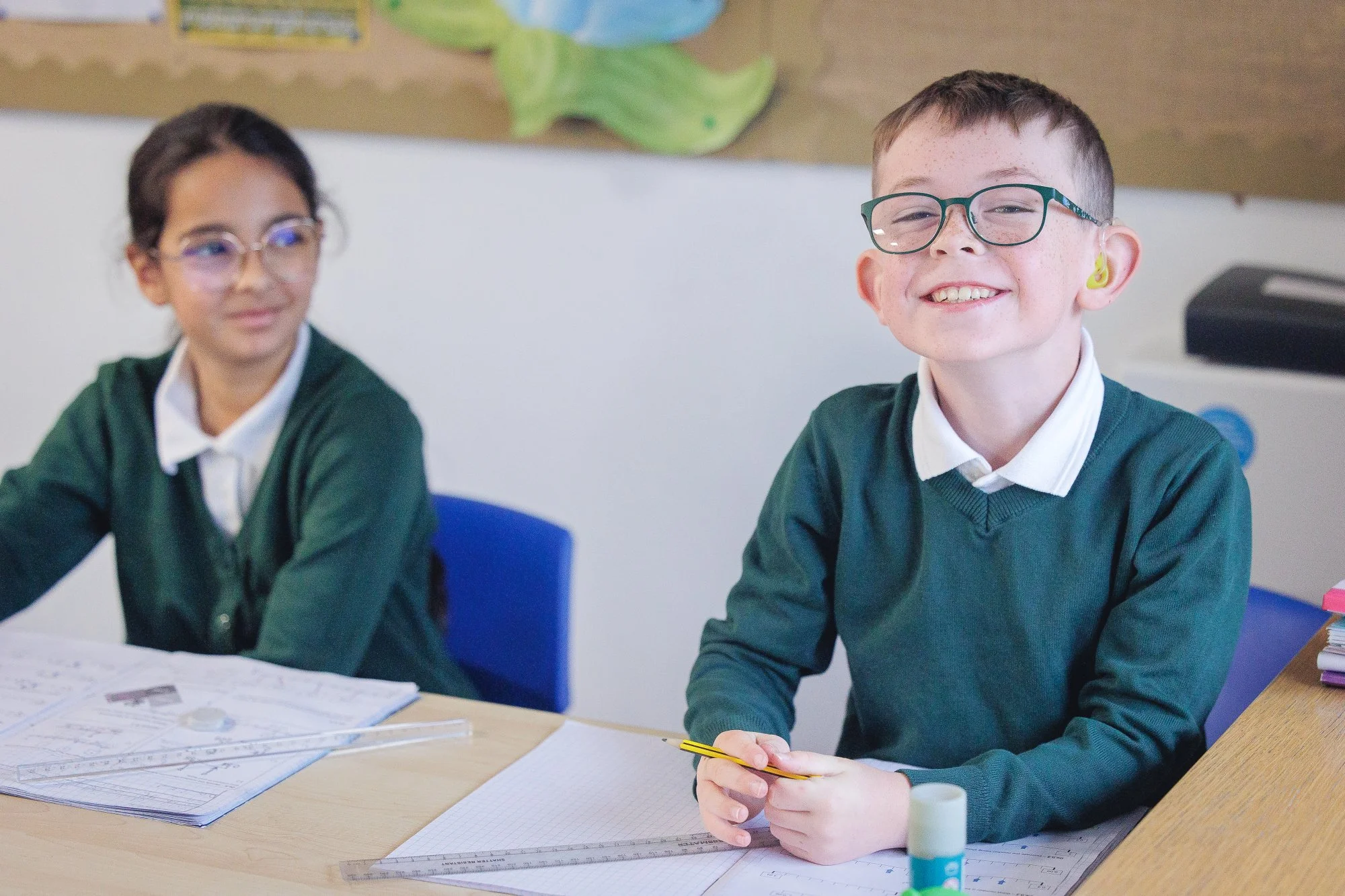 Smiling boy with glasses and hearing aids sitting at a desk in a classroom, holding a yellow pencil. A girl with glasses is sitting next to him, with papers, a ruler, and school supplies on the desk.