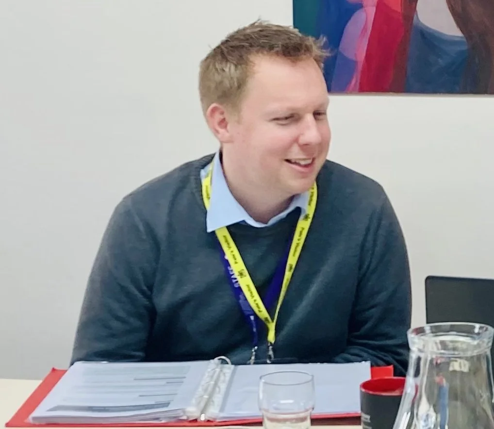 A man sitting at a table, smiling, wearing a blue shirt with a dark sweater, with a yellow and blue lanyard around his neck, in an office setting with a white wall and colorful artwork in the background, and various items on the table including a red binder, glass of water, and a pitcher.