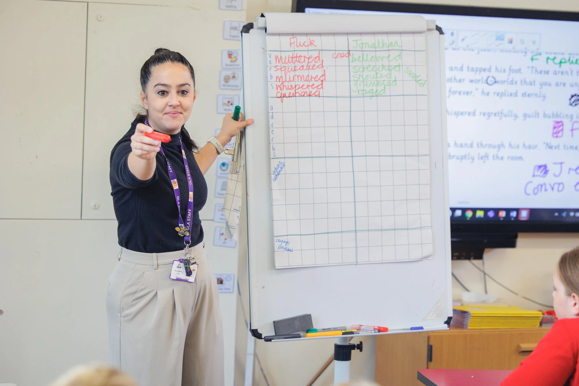 A woman with dark hair in a ponytail, wearing a black long-sleeve top and beige pants, is standing and pointing at a whiteboard with a red marker. The whiteboard contains a list of words written in red, green, and blue markers, divided into columns. There are students sitting in front of her, with at least one visible wearing a red shirt. A large screen behind her displays typed text annotations.