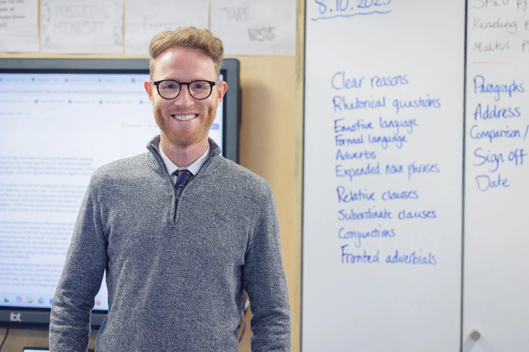 A smiling man with glasses and a gray sweater stands in front of a whiteboard filled with written notes, with a computer monitor behind him in a classroom or office setting.