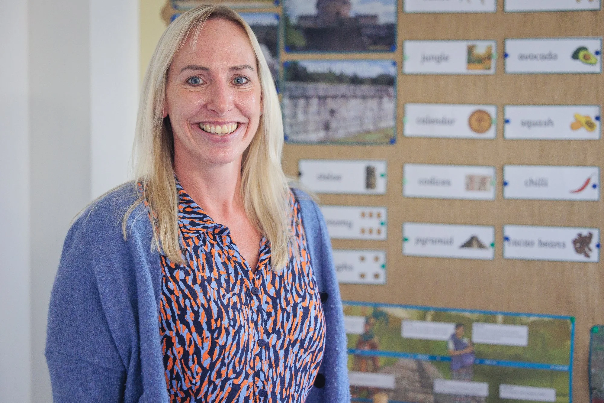 A smiling blonde woman wearing a patterned shirt and a blue cardigan stands in front of a bulletin board with world history images and captions.