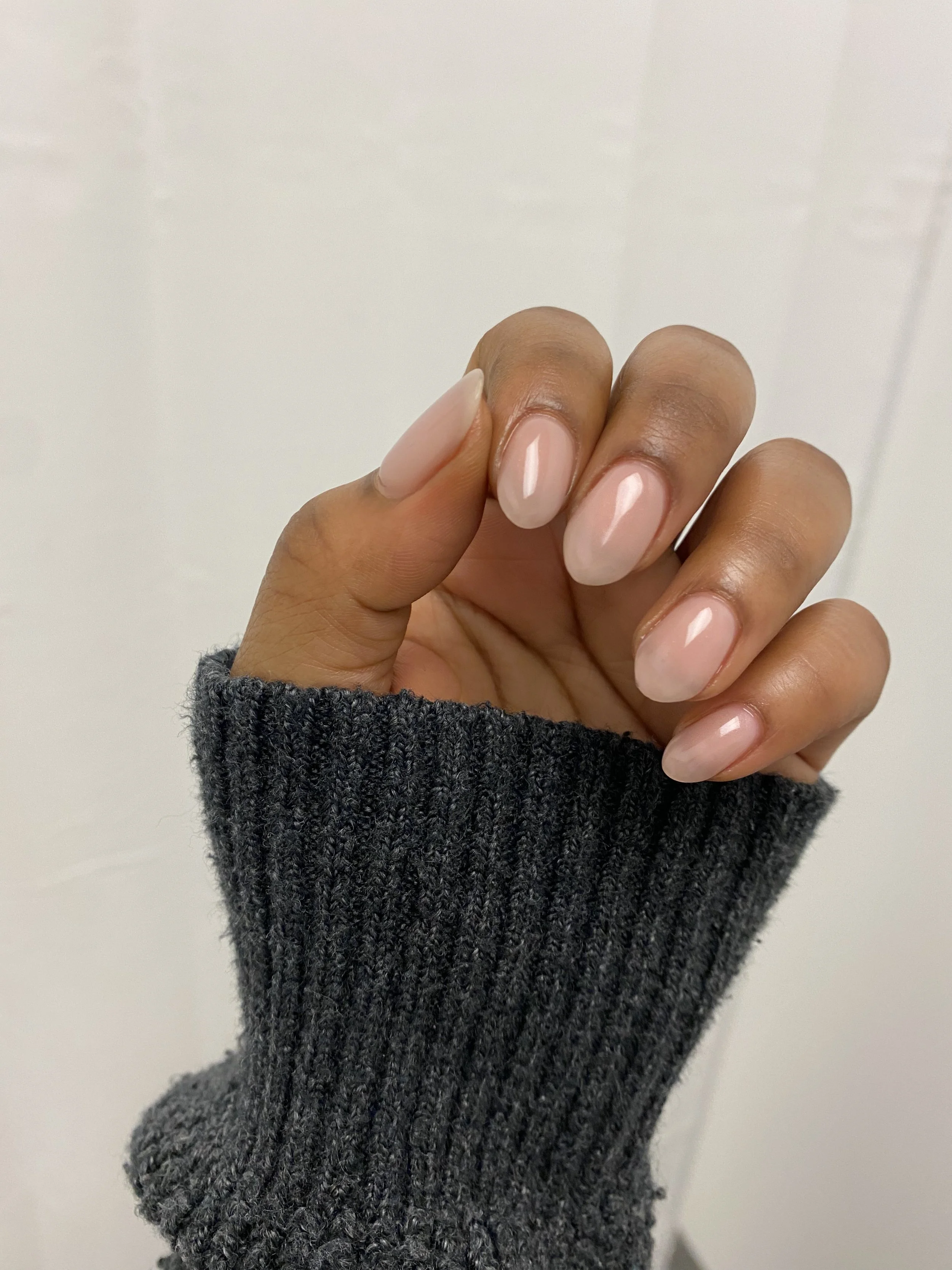 Close-up of a hand with neatly manicured, shiny, light pink nails. The hand is partially covered by a dark gray, ribbed sweater sleeve. The background is plain and white. all done by Acclaim Nailz gentle gel nail services.