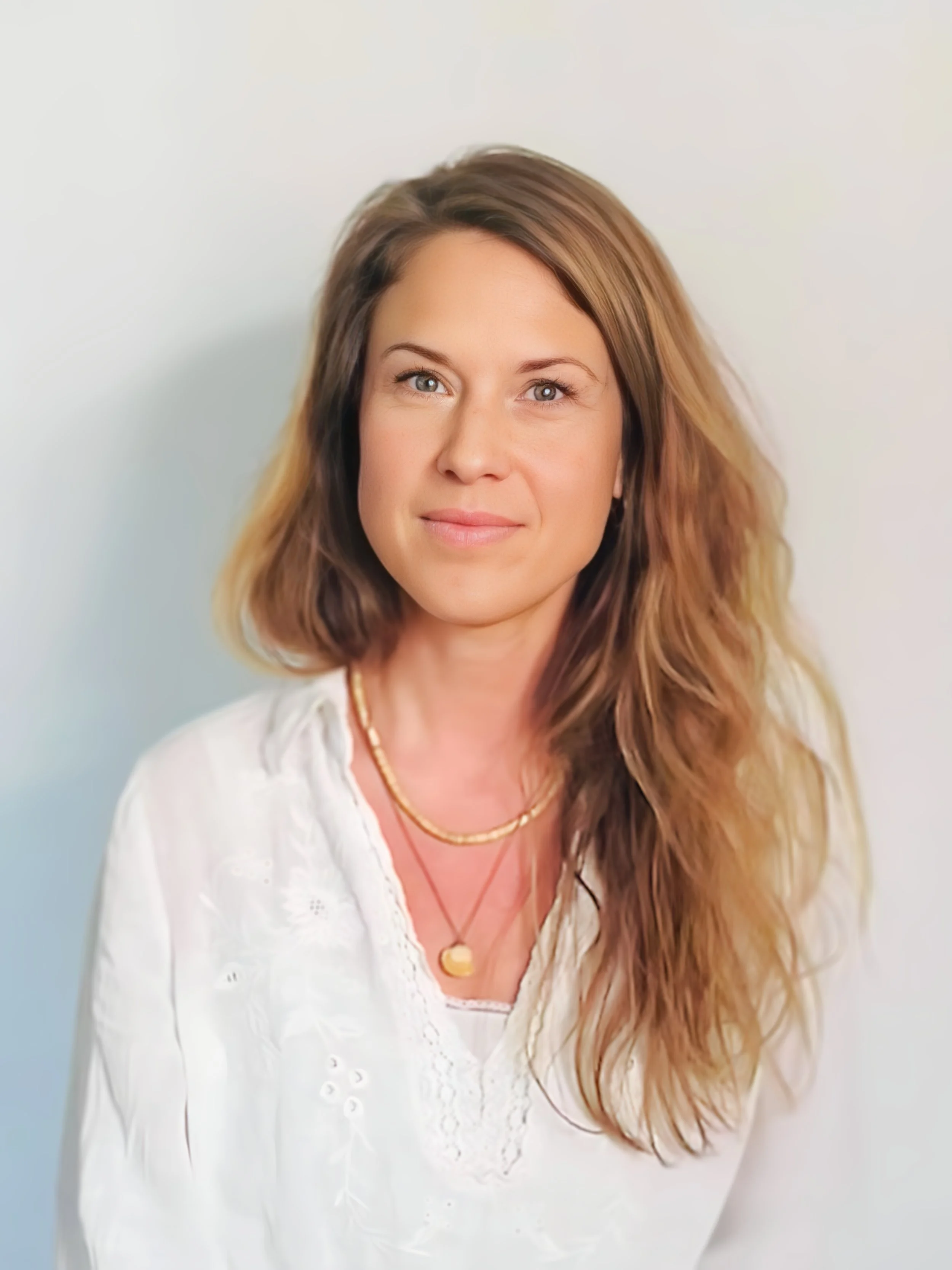 Porträt einer Frau mit langen braunen, lockigen Haaren, blauer Augen, in weißem Spitzentop und Goldschmuck vor weißem Hintergrund.