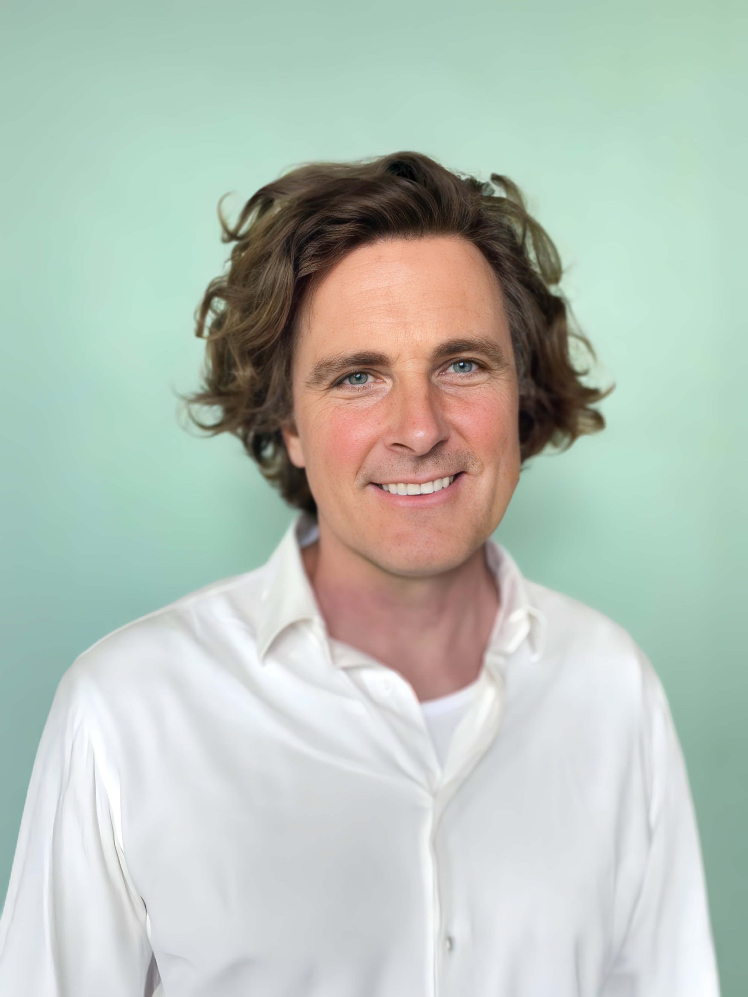 Porträt eines lächelnden Mannes mit mittellangen, lockigen braunen Haaren, blauen Augen, weißen Zähnen, trägt ein weißes Hemd, vor einem hellgrünen Hintergrund.