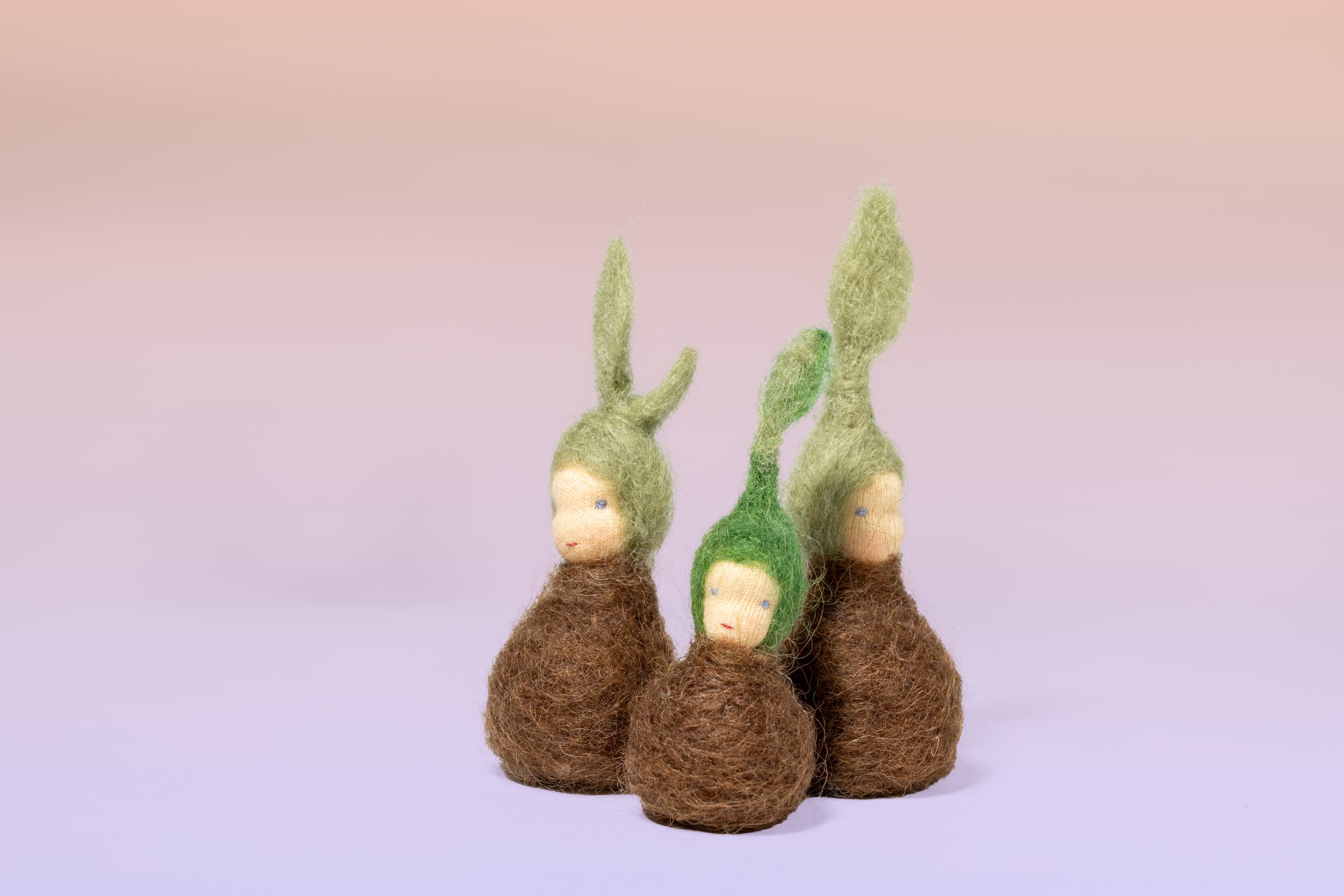 Drei Wollfiguren in Form von kleinen Wesen mit bunten Hüten in Form von Blättern die an Pflanzen oder Pilze erinnern, vor einem pastellfarbenen Hintergrund.