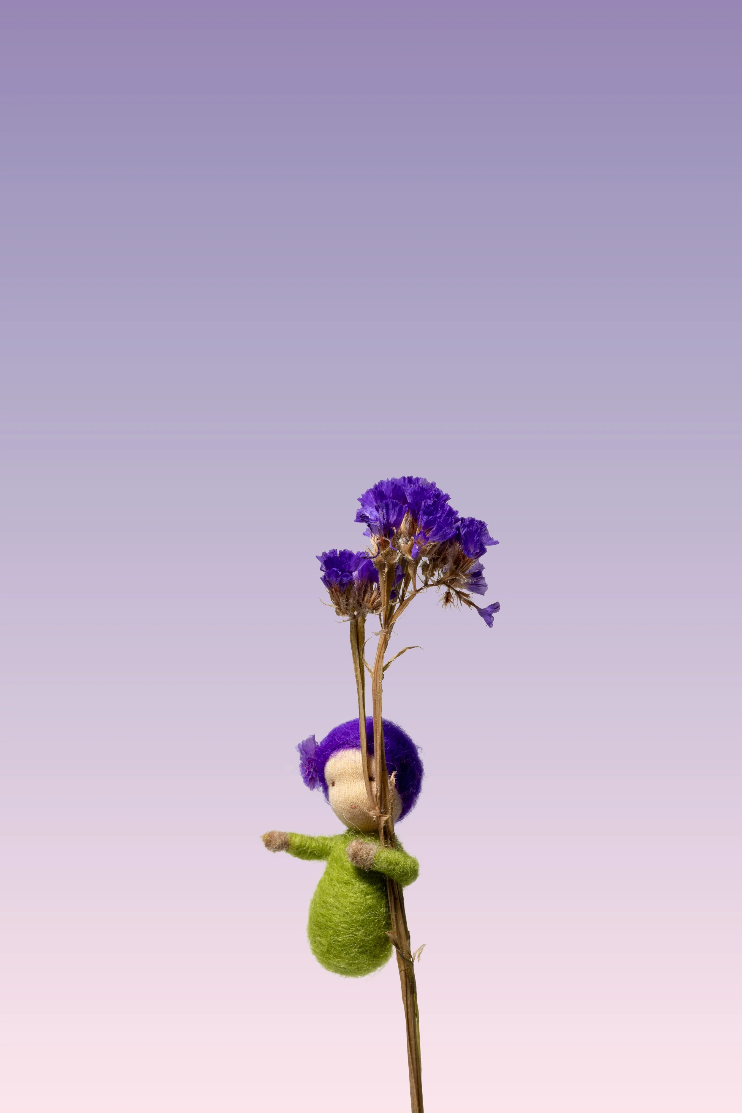 Kleine Filzpuppe mit lila Haar, die an einem Stängel mit blauen Blumen hängt.