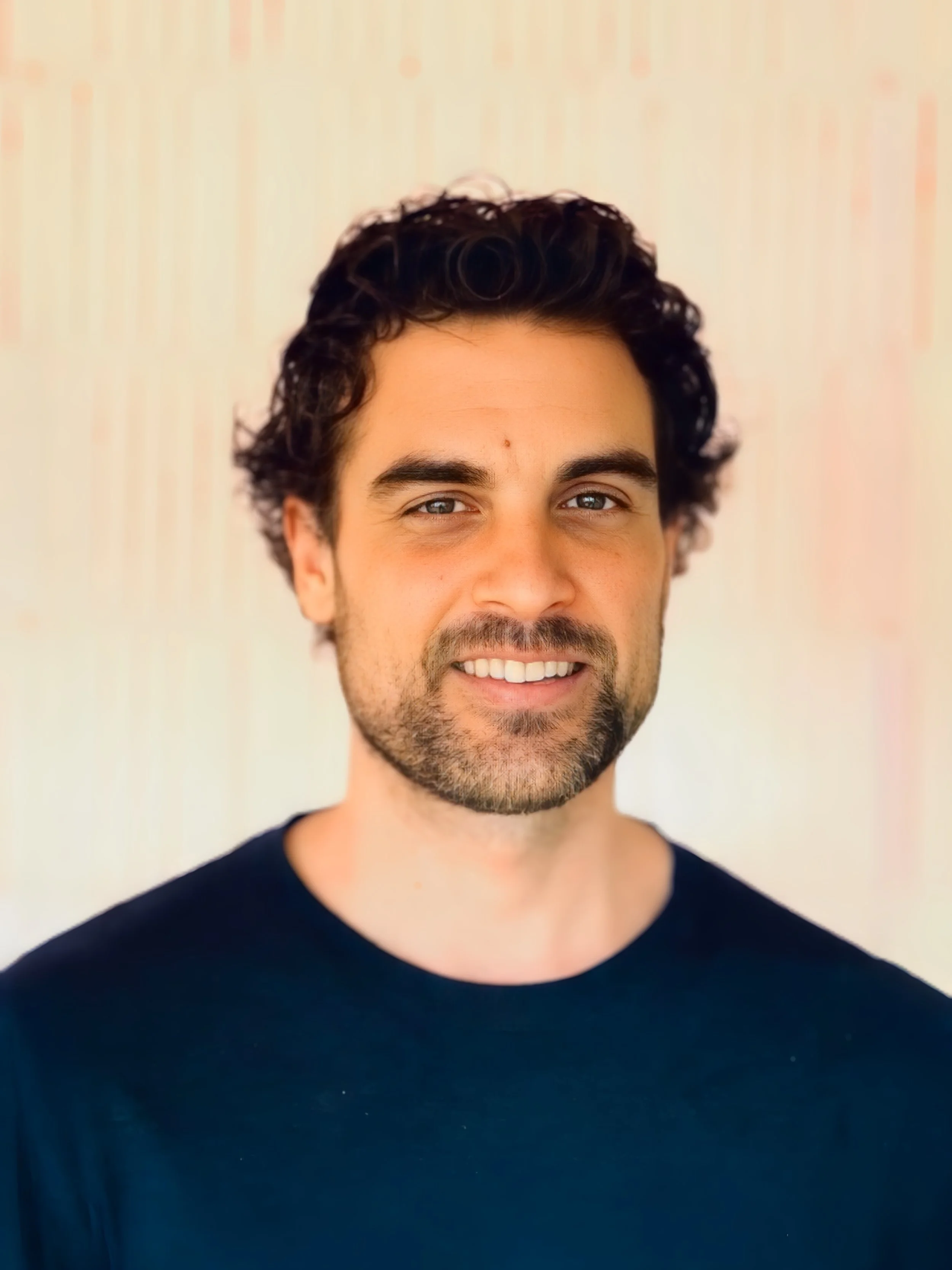 Mann mit braunen, welligen Haaren, Bart und brauen Augen, lächelnd, trägt ein schwarzes T-Shirt