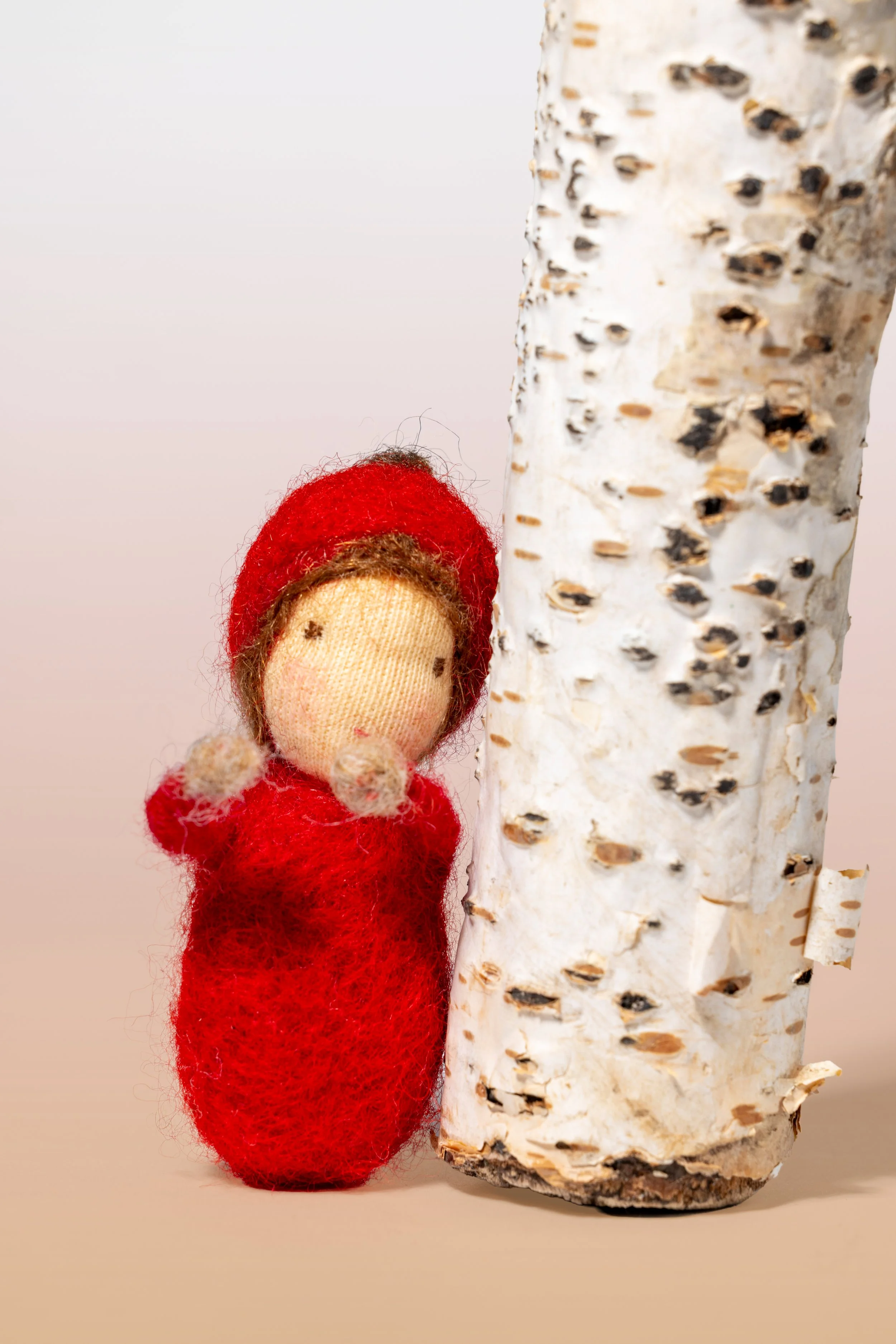 kleine handgefertigte Filzfigur in rotem Kleid und rotem Hut steht neben einem Baumstamm, Jahreszeitenpüppchen,nach Waldorf-Art.