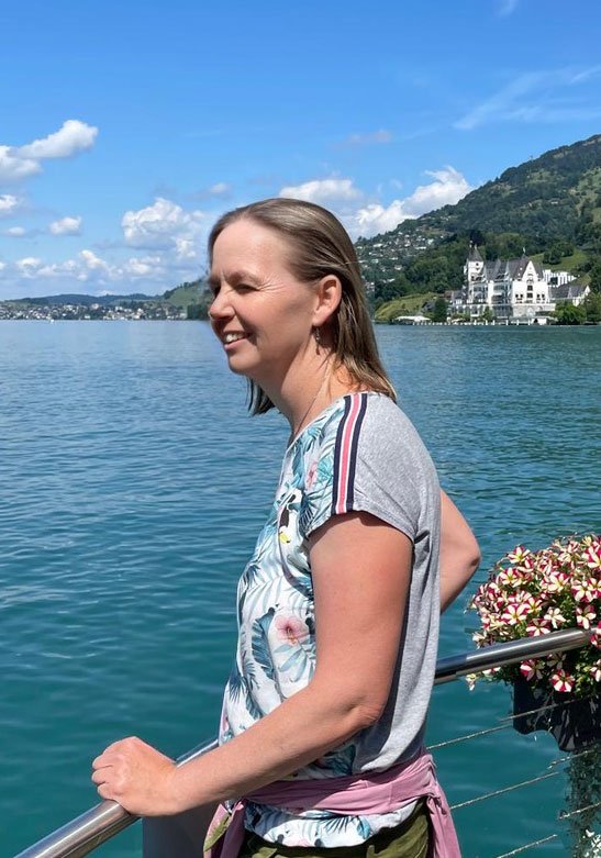 Frau am Ufer eines Sees, lachend, im Hintergrund Berge und ein Schloss, sonniger Tag.