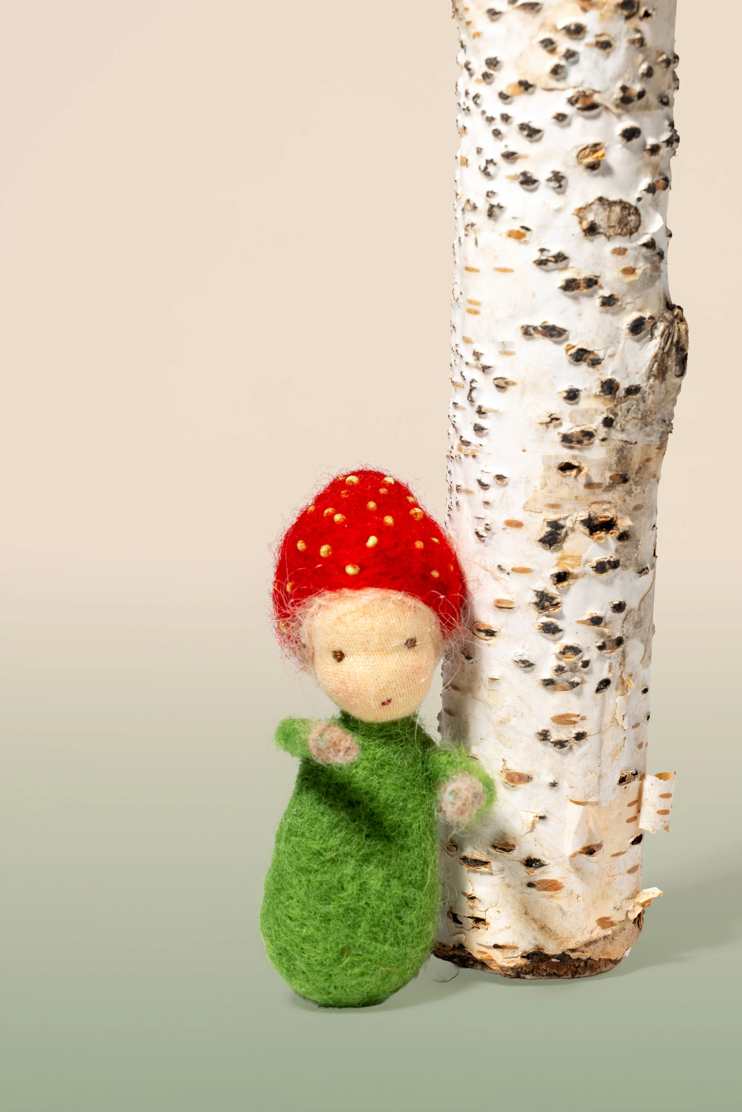 Eine kleine handgefertigte Filzfigur mit roter Mütze, die wie eine Erdbeere aussieht, steht neben einem Birkenstamm auf neutralem Hintergrund.
Jahreszeitenpüppchen,nach Waldorf-Art