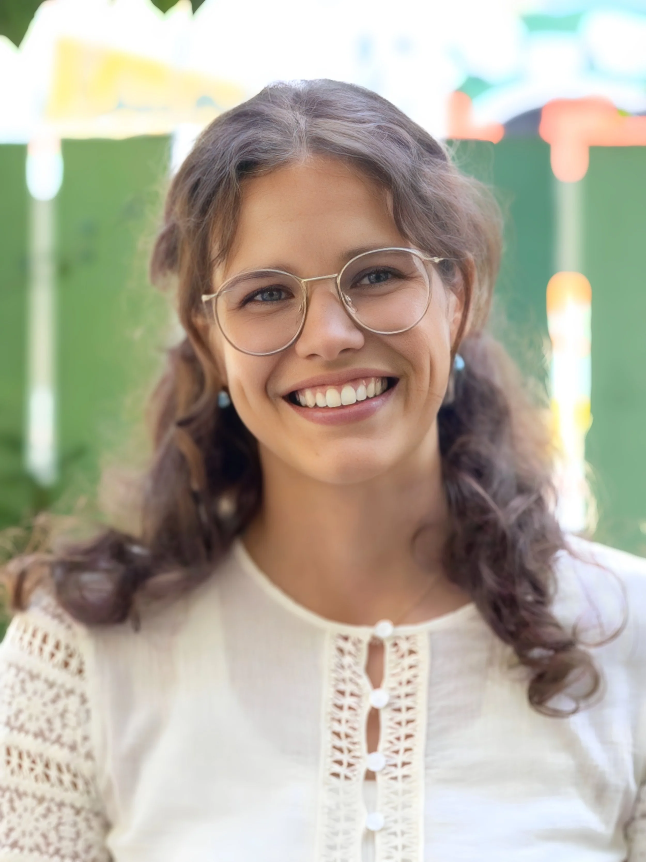 Lächelnde junge Frau mit Brille, braune lockige Haare, weiße Bluse, im Garten.