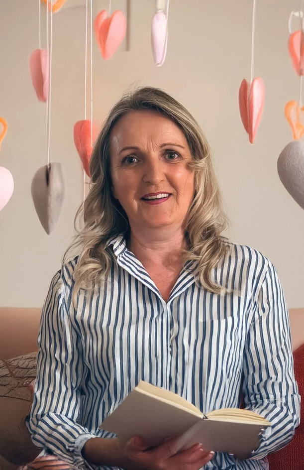 Eine Frau mit blonden, lockigen Haaren sitzt auf einem Sofa vor einer Wand, die mit hängenden Herzen in verschiedenen Pastellfarben geschmückt ist, und liest ein offenes Buch.