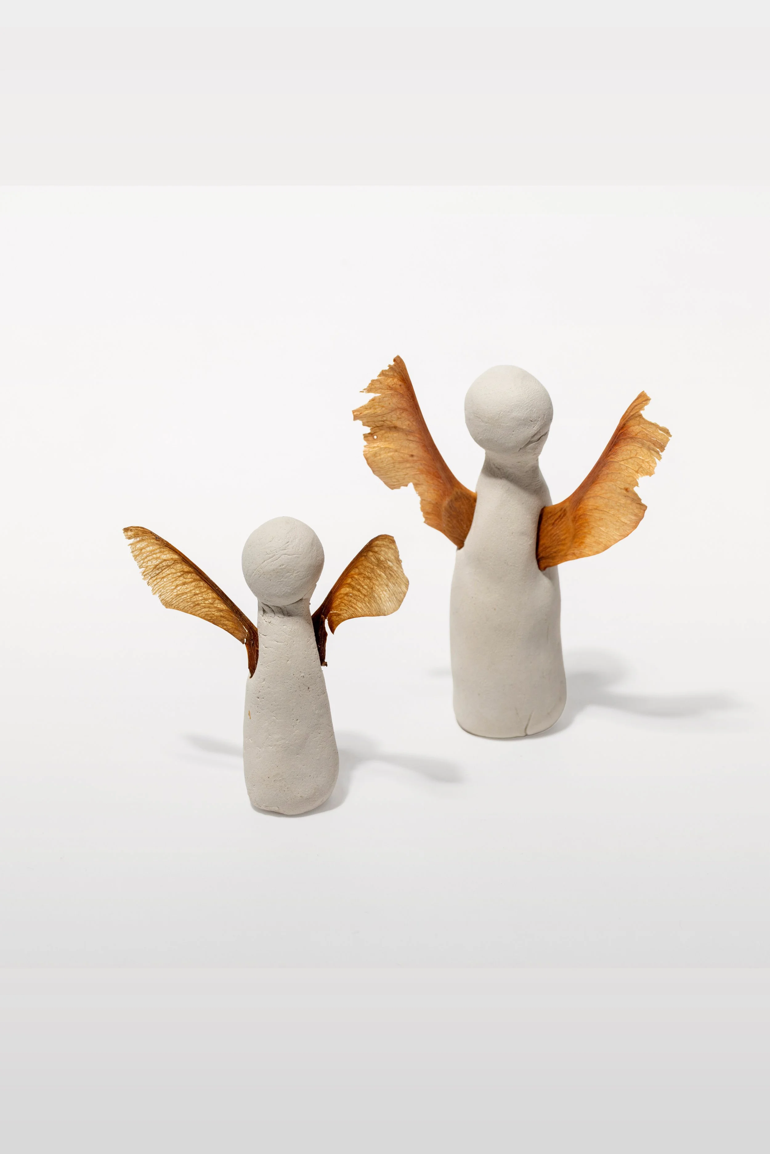 Zwei Engel-Dekorationen aus weißem Material mit braunen getrockneten Blättern als Flügel, unterschiedlicher Größe, auf weißem Hintergrund.