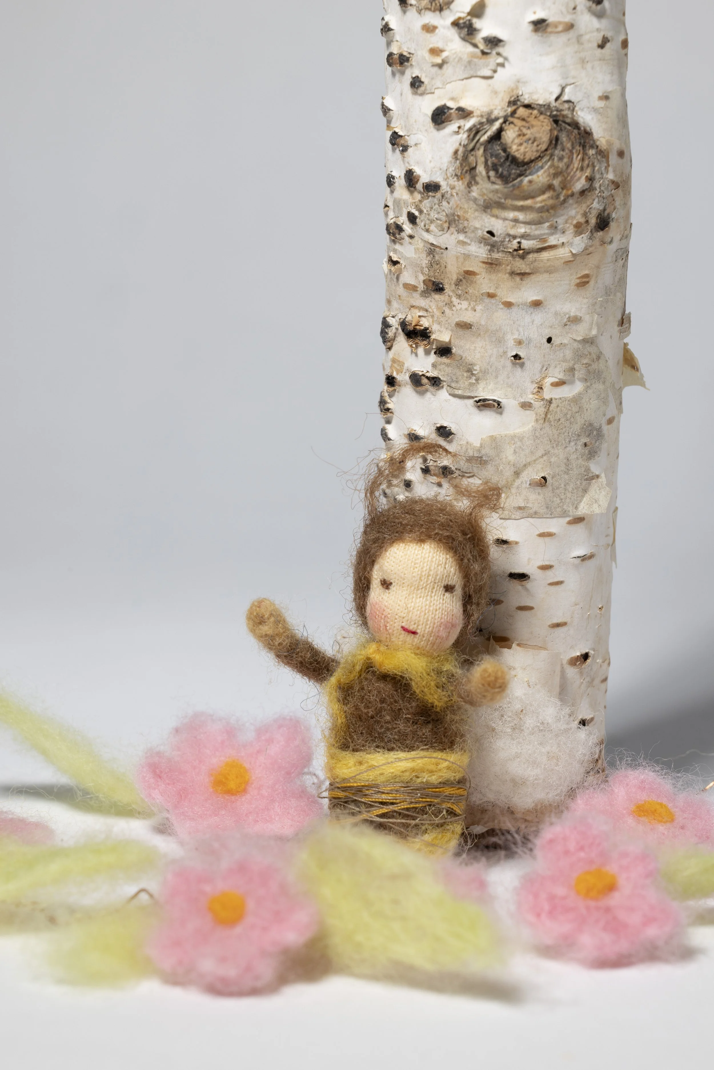 Kleine Filzfigur Biene nach Waldorf-Art aus hochwertiger Schurwolle handgefertigt vor einem Birkenstamm, umgeben von Filzblumen in pink und grün.