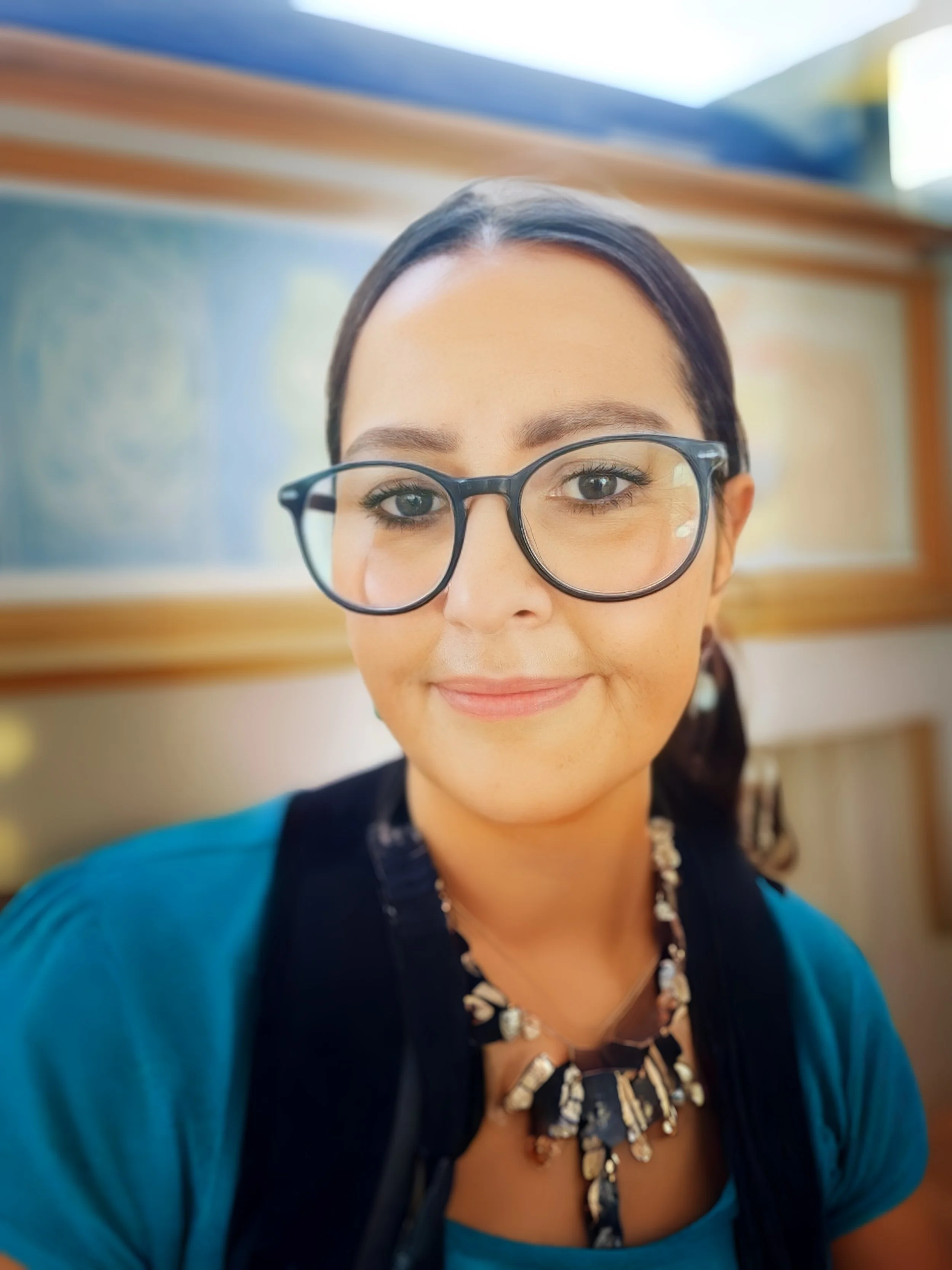 Nahaufnahme eines lachenden Frauenkopfes mit schwarzer Brille, braunen Haaren, blauer Bluse und einer Kette, im Hintergrund eine beschriftete Tafel.