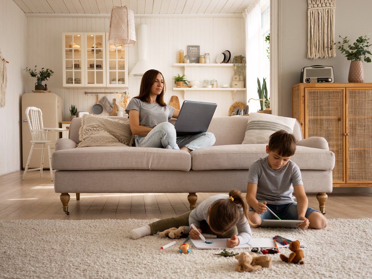 A woman sitting on a beige couch working on a laptop, two children playing on the floor with toys and drawing on paper, in a bright, cozy living room with white walls, wooden furniture, and decorative plants.