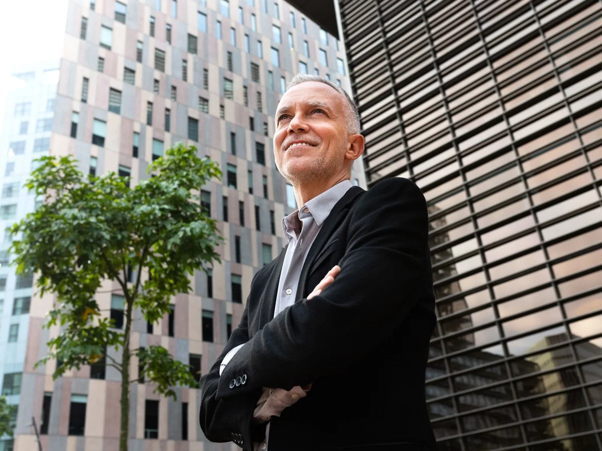 A confident middle-aged man in a black blazer and gray shirt standing outdoors in an urban setting with tall office buildings and a green tree behind him.