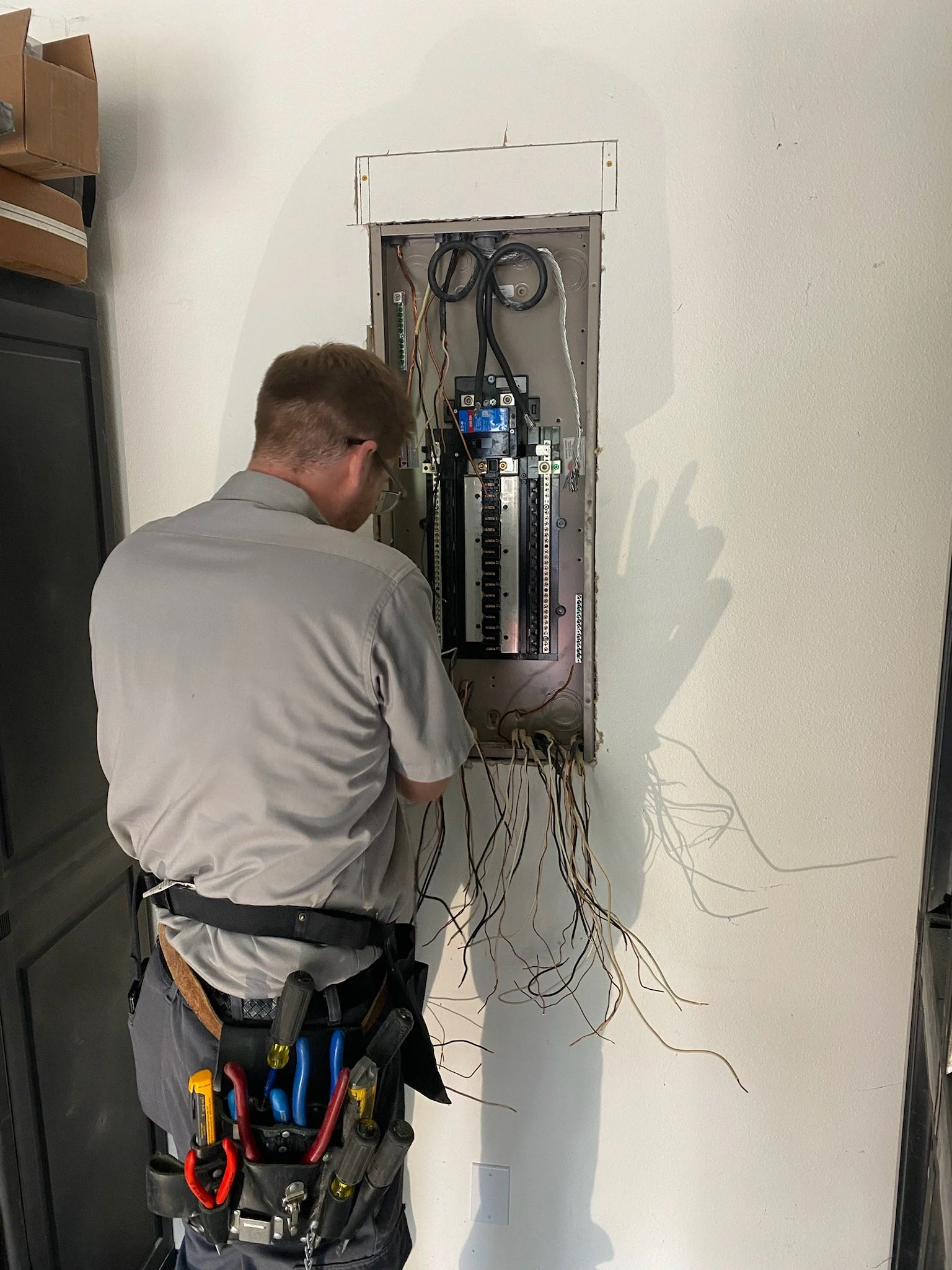 A technician working on an electrical panel with exposed wiring, standing in front of a white wall.