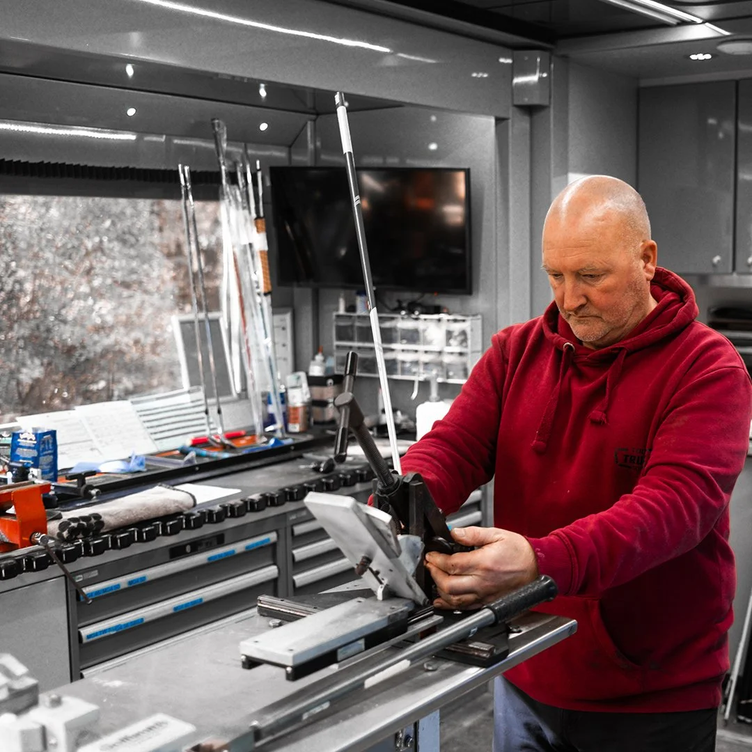A man in a TaylorMade cap and blue shirt working on golf club equipment in a modern workshop.