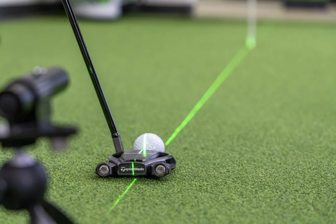 Golf ball on a tee with a golf club hitting it, and a green laser line on the grass