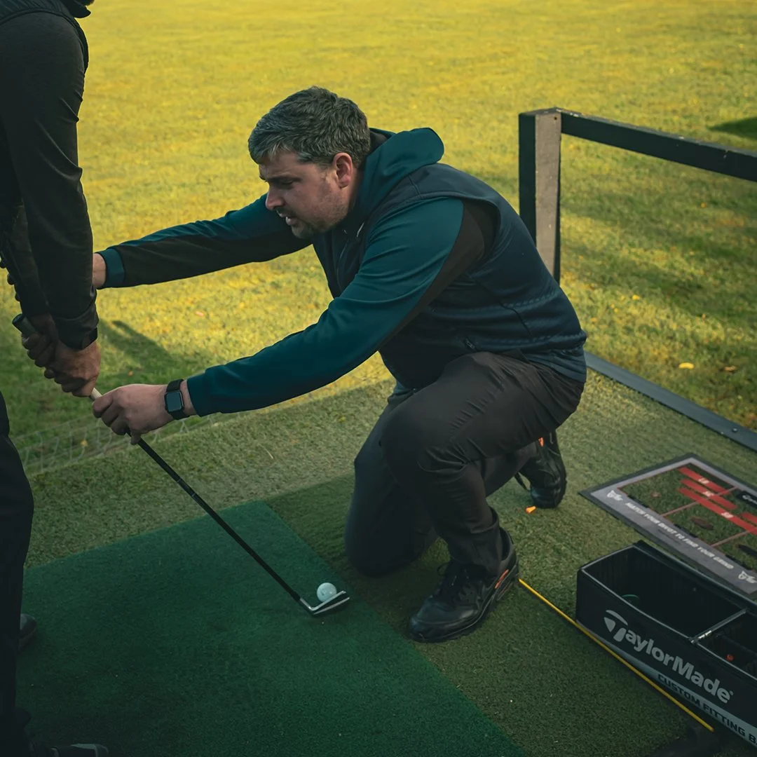 A person playing golf on a golf course during sunset, swinging a club with a golf ball in mid-air, golf bag and equipment nearby.