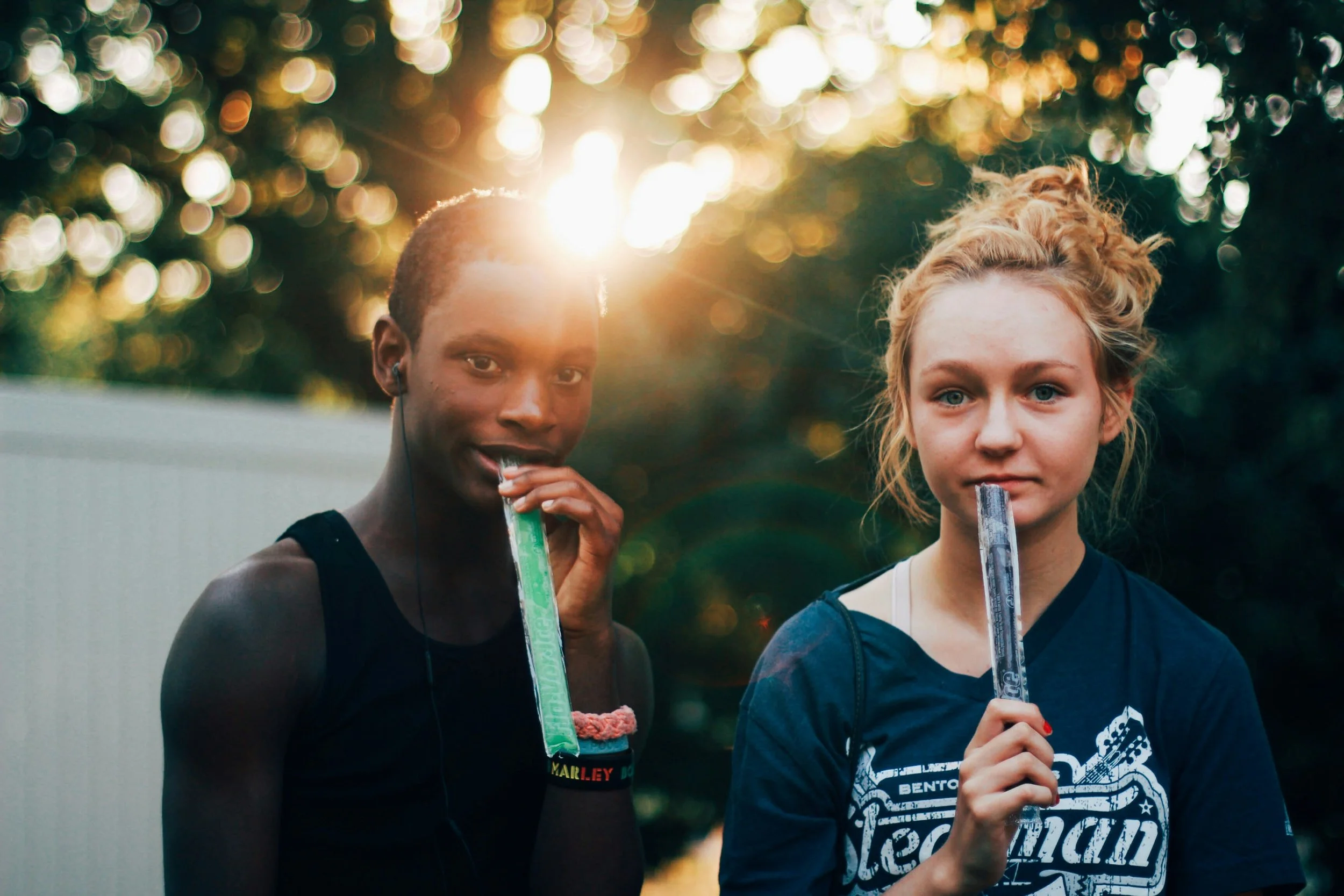 A boy and a girl standing outdoors with sunlight behind them, eating popsicles.