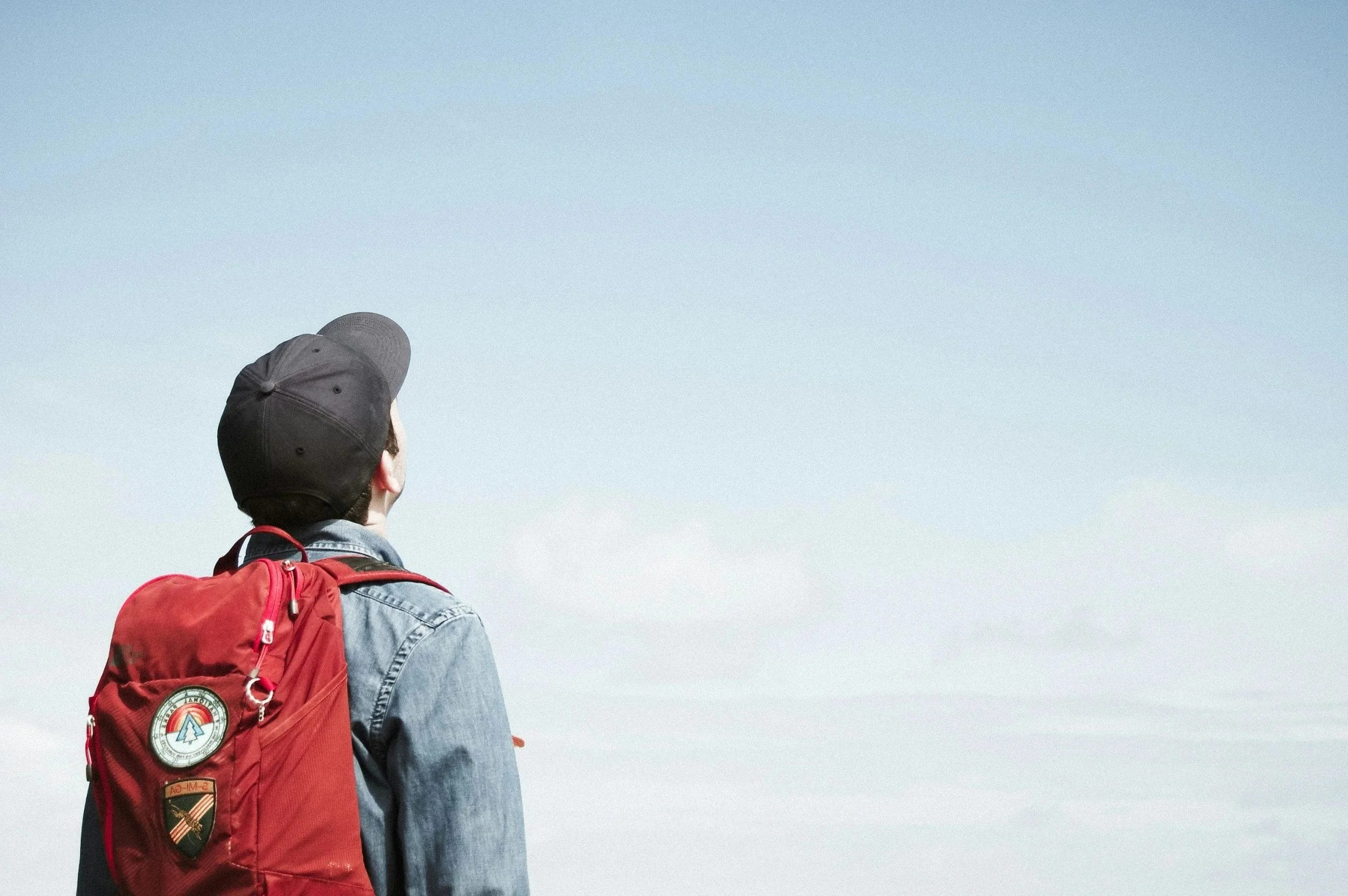 A person wearing a black cap and denim jacket with patches, carrying a red backpack, looks up at the sky.