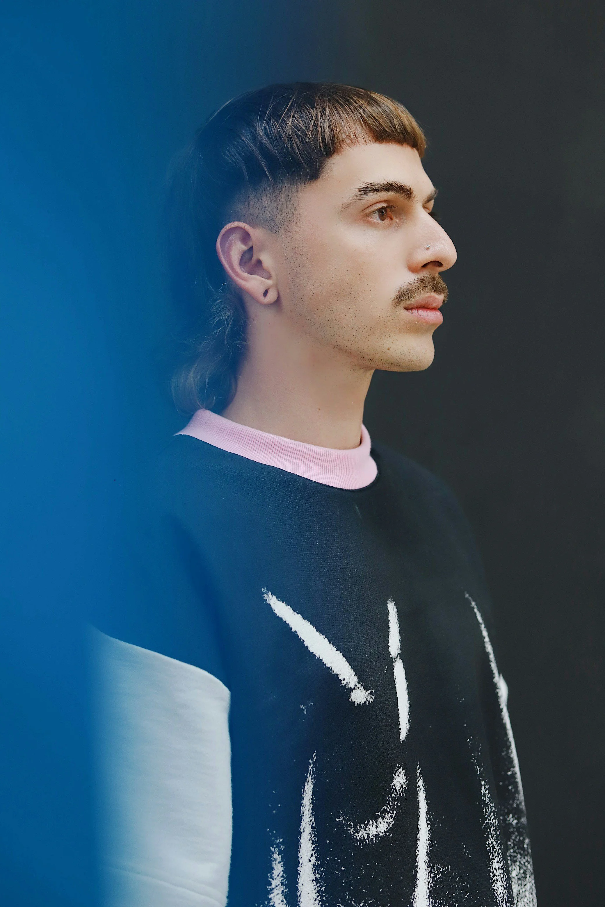 A young man with a mustache and a nostril piercing, looking to the right, wearing a black and white sweater with a pink collar and a dark background