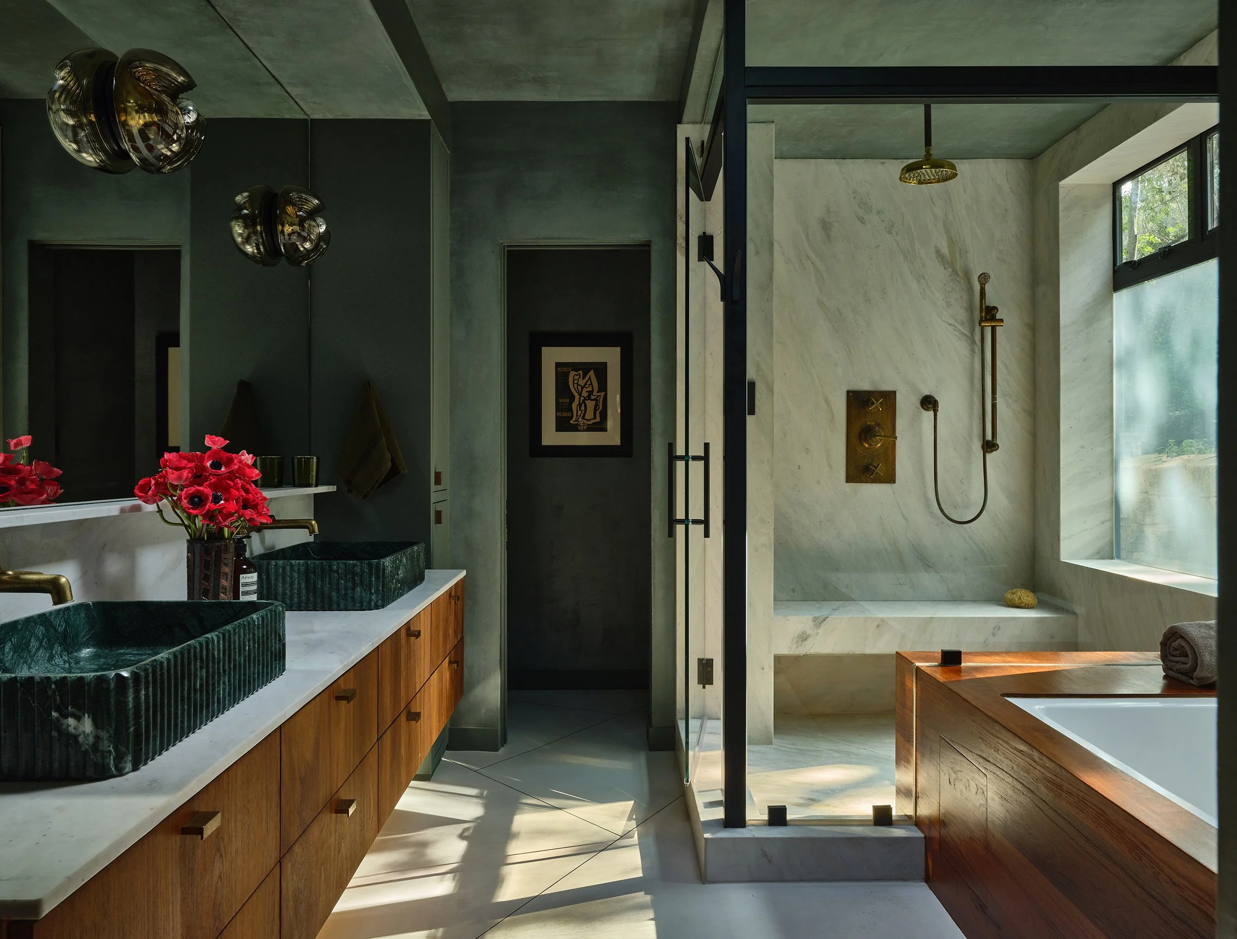Anthony Jeselnik’s primary bathroom room, interior design by Riccio Vidaurre Design – A dark and moody bathroom with grey green plaster walls, green stone sinks and a wooden bathtub. A white marble shower with brass fixtures and a glass enclosure.