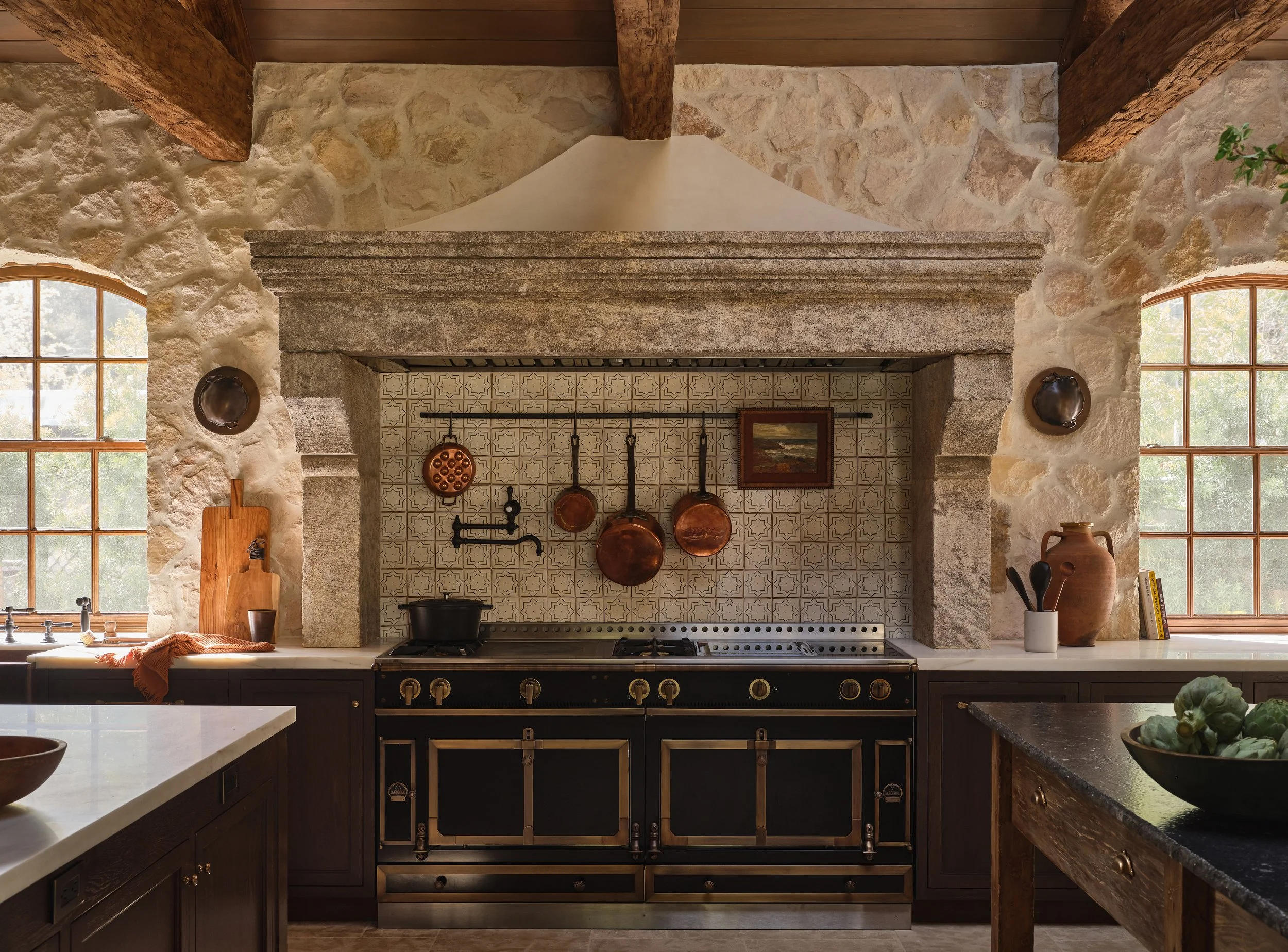 Rustic kitchen with stone walls, wooden beams, a vintage black stove, copper pots hanging above, and large windows with a view of trees outside.