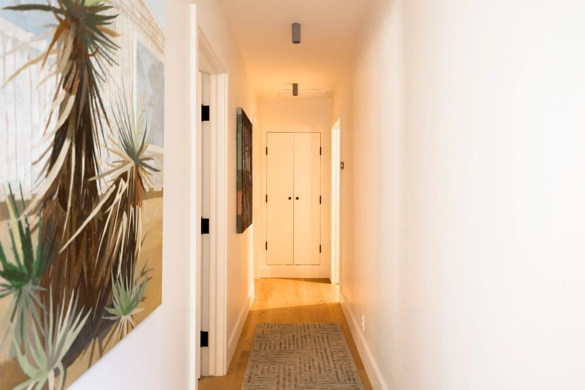A hallway in a mid century modern home, with white walls, wood oak floors and grey surface mount can lights.  Large paintings of foliage cover the walls.