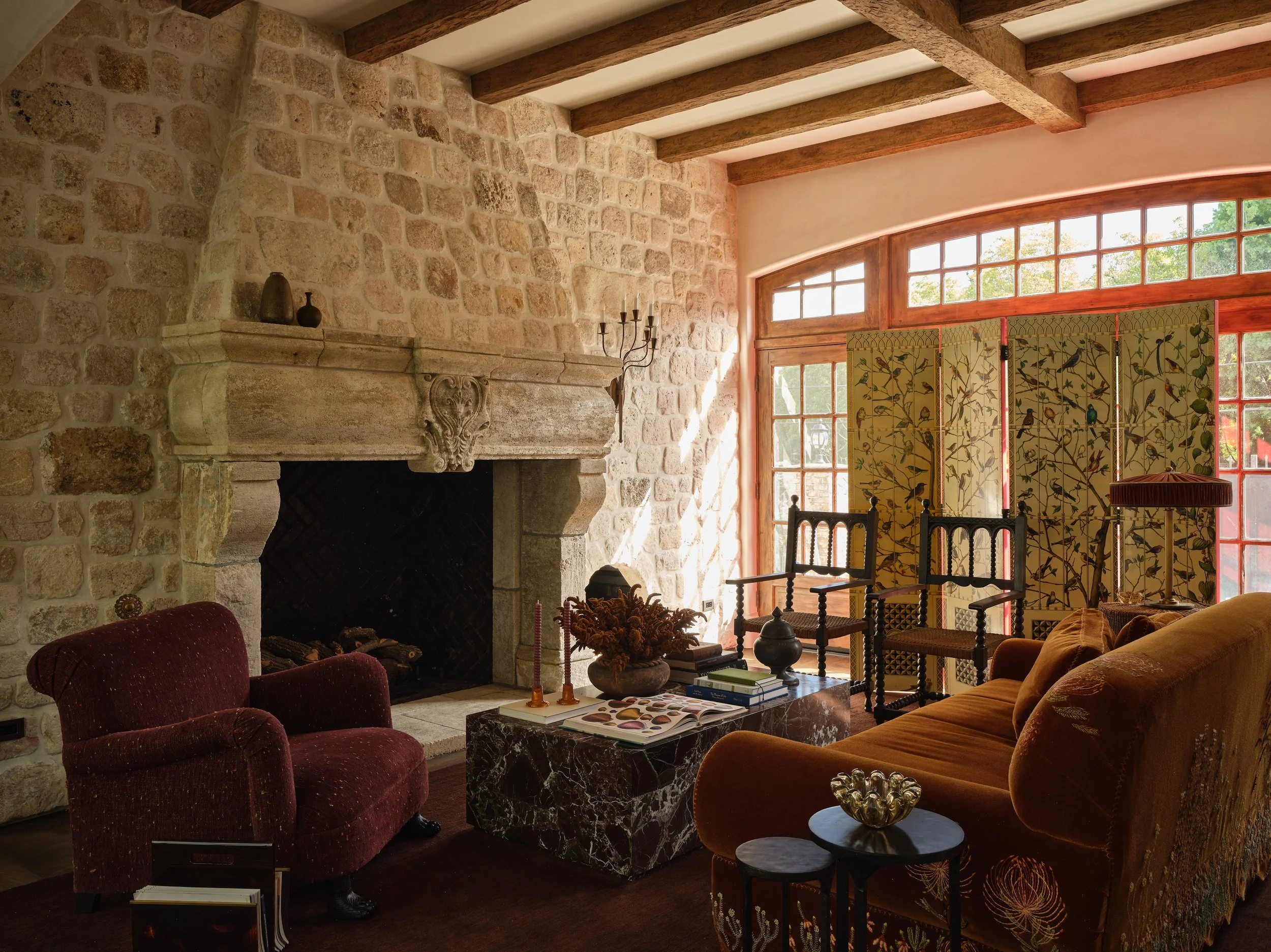Living room with stone fireplace, large window, wooden ceiling beams, antique chairs, velvet sofa, marble coffee table, and decorative folding screen.