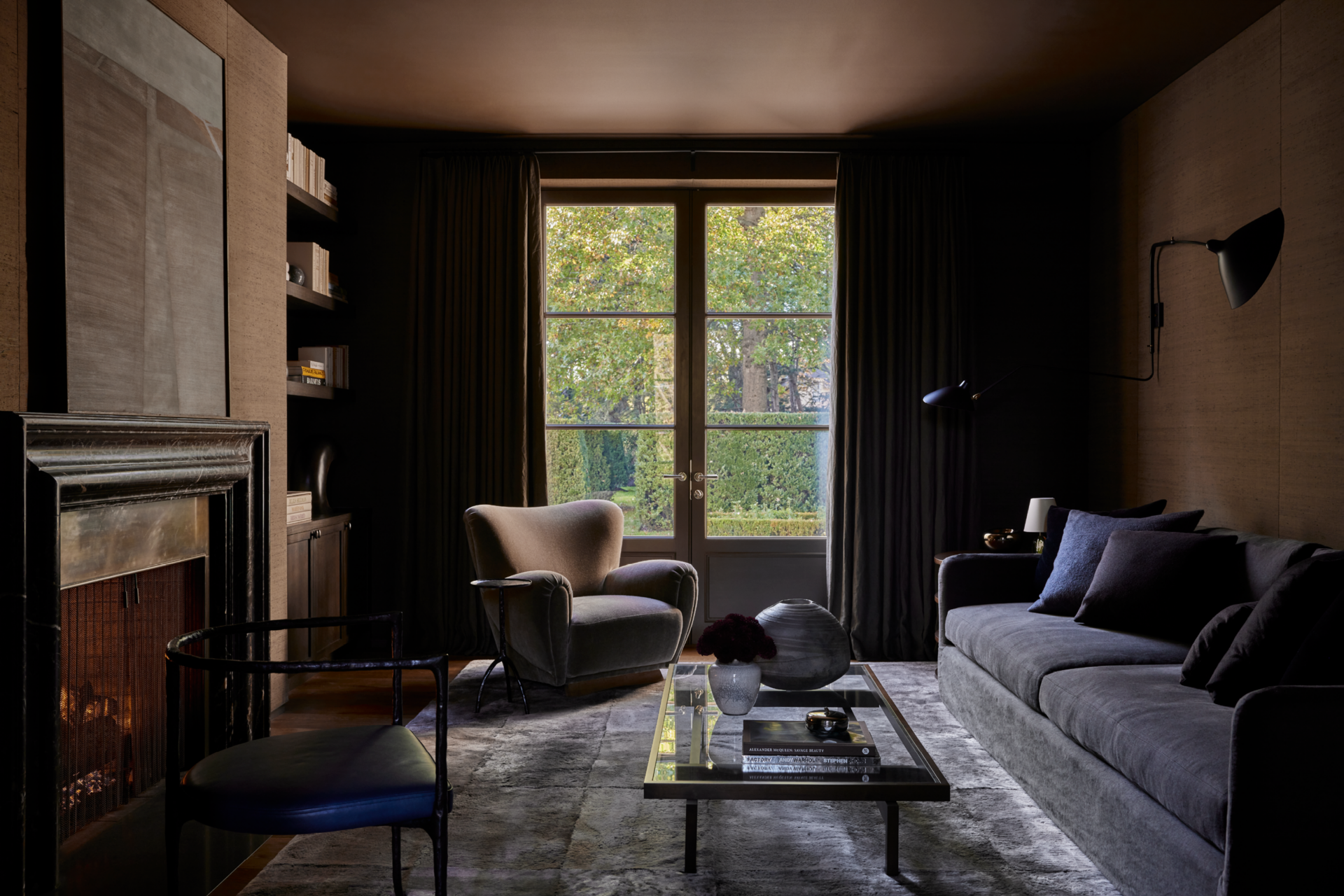 A dimly lit living room with a large window showing green trees outside, a gray sofa with black pillows, a beige armchair, a glass coffee table with decorative items and books, dark curtains, a fireplace, and modern wall-mounted lamps.