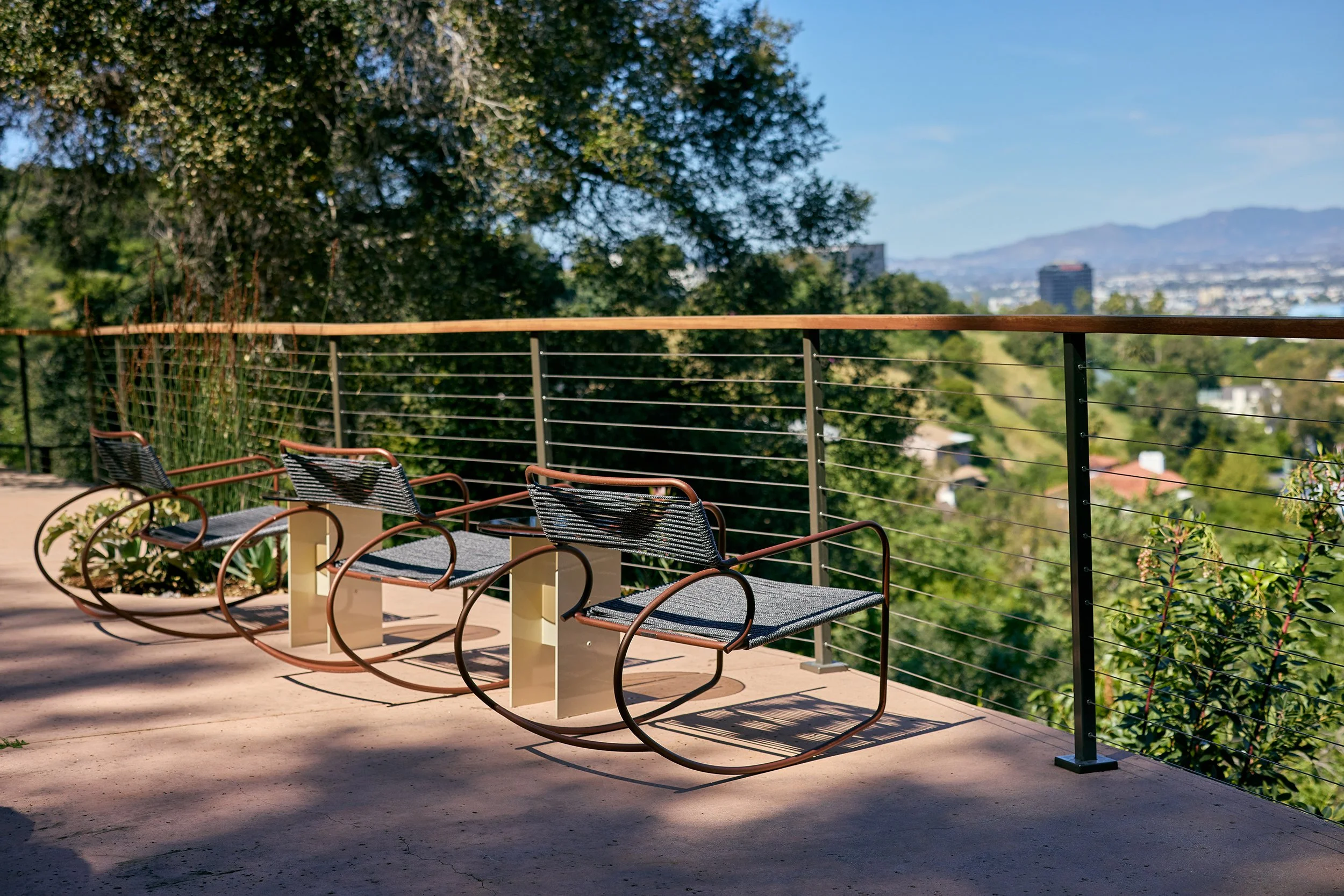 Anthony Jeselnik’s back yard, interior design by Riccio Vidaurre Design – row of Walter Lamb Rocking Chairs by Brown Jordan sit beside “Rugosa” side tables by Kalon Studios overlooking a view of los angeles.