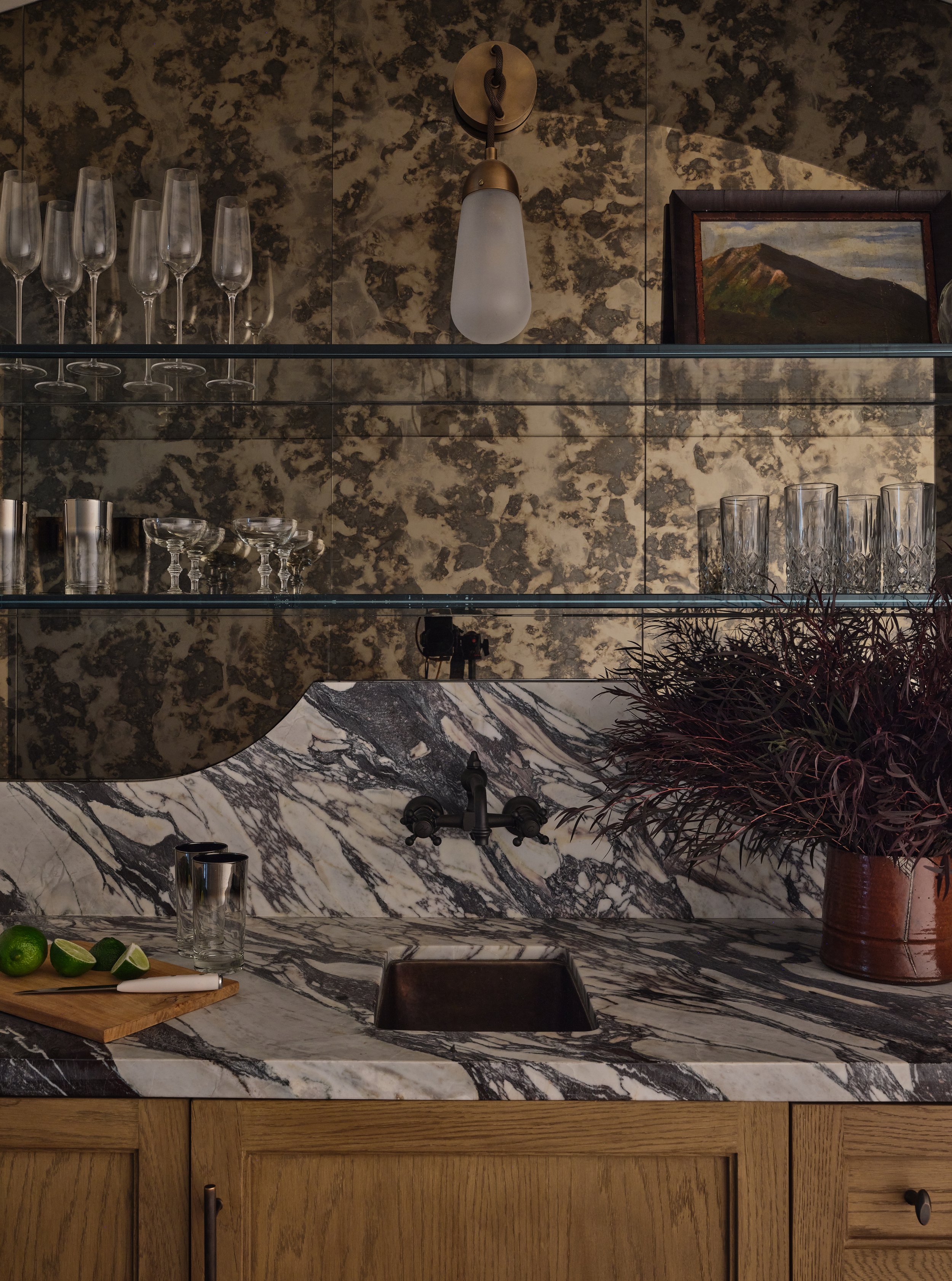 A kitchen with a marble countertop and backsplash, a black faucet, top shelves with glassware, a wooden cabinet, a potted plant, and a wall-mounted light fixture